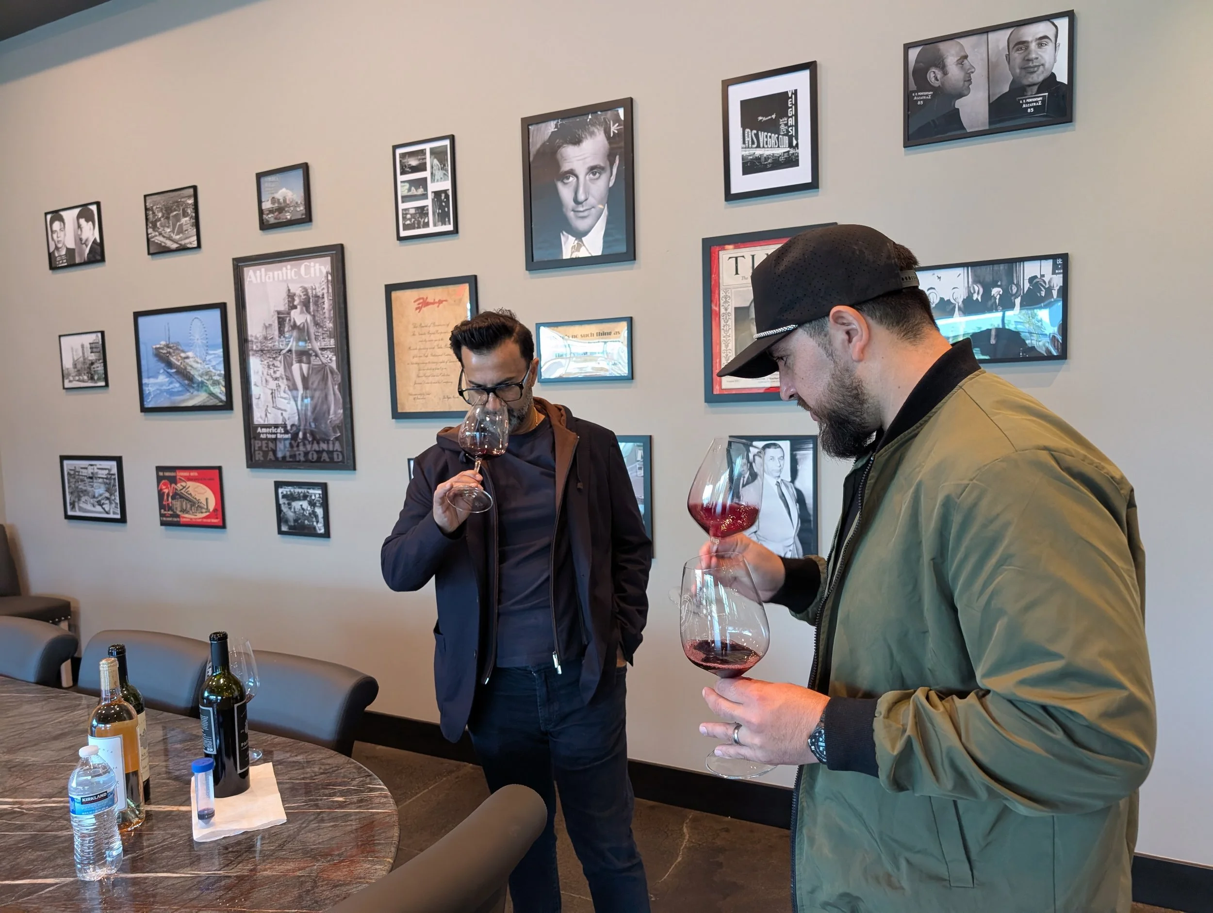 Two men tasting red wine in a room with a gallery wall of black-and-white photographs and artwork.