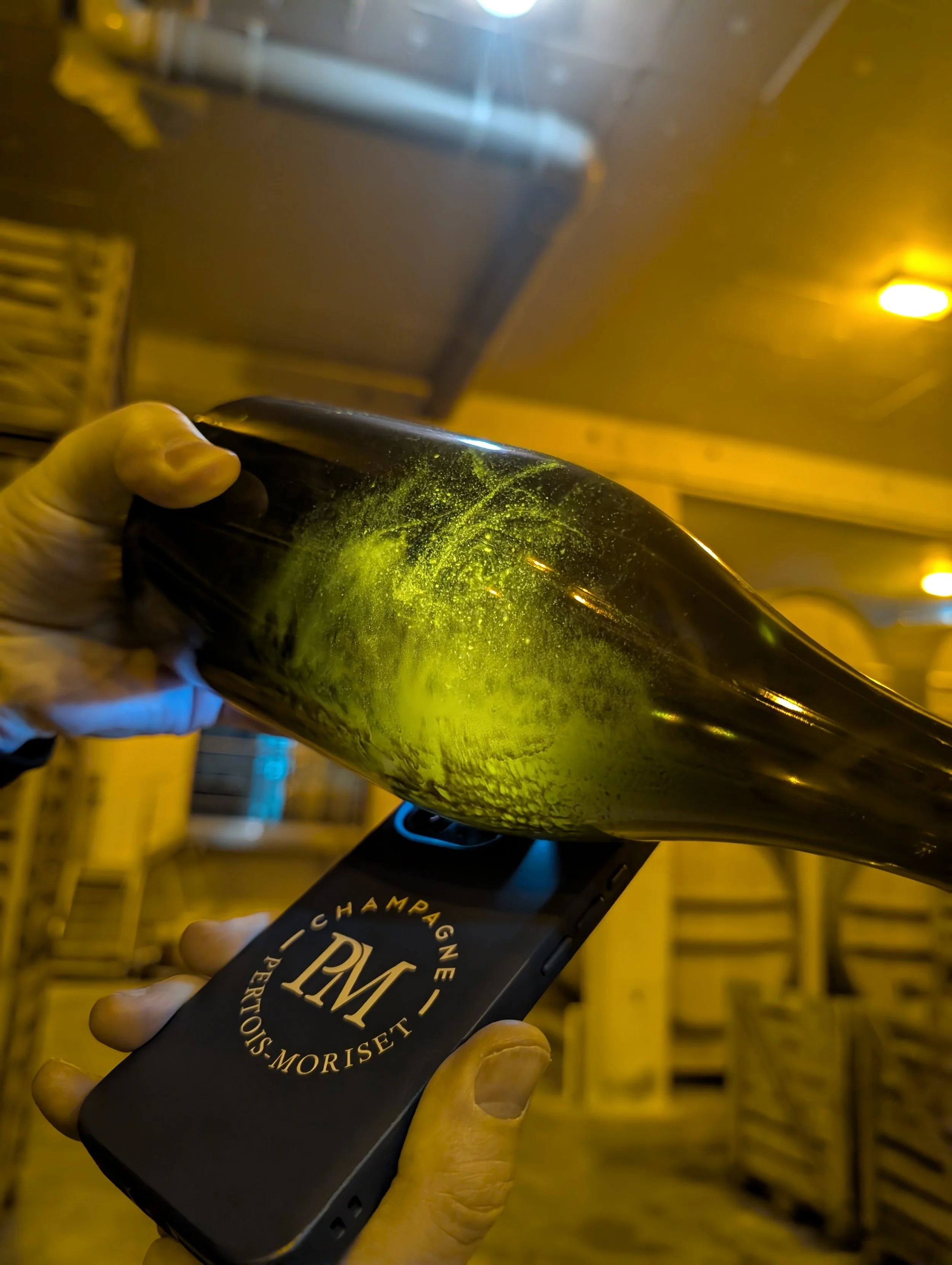 Person holding a champagne bottle and a phone with a logo reading 'Champagne Moriset'. The bottle appears to be leaking greenish liquid.