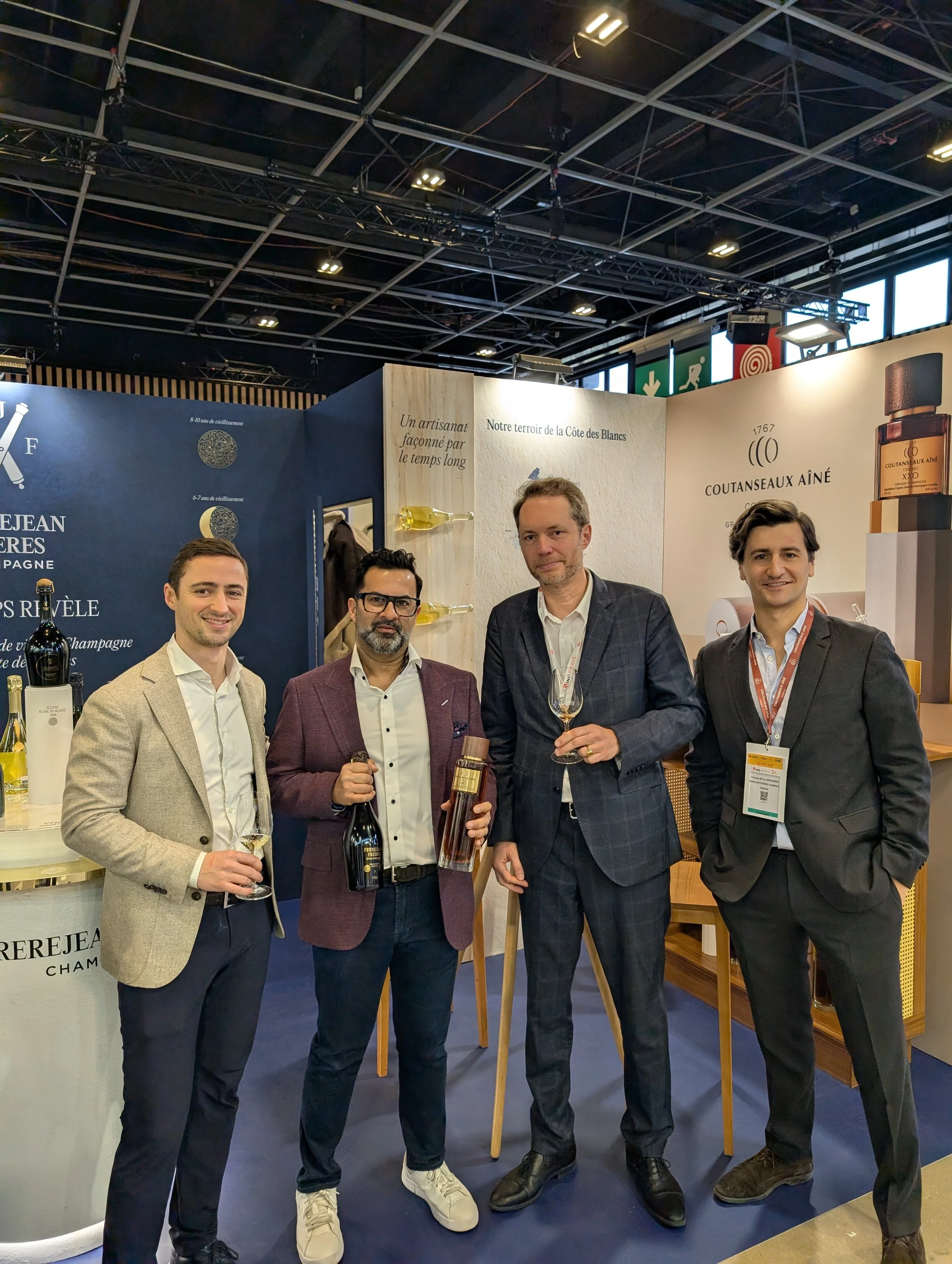 Four men in suits standing together at a trade show booth, holding wine bottles and glasses, with promotional banners and bottles of champagne in the background.