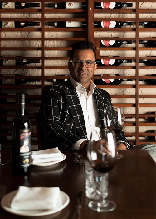 Man in a checkered blazer and white shirt holding a glass of red wine at a restaurant table.