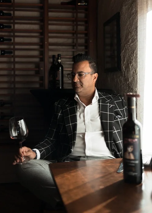 A man in a checkered blazer and white shirt sitting at a table in a restaurant, holding a glass of red wine, with a bottle of wine and a window with curtains nearby.