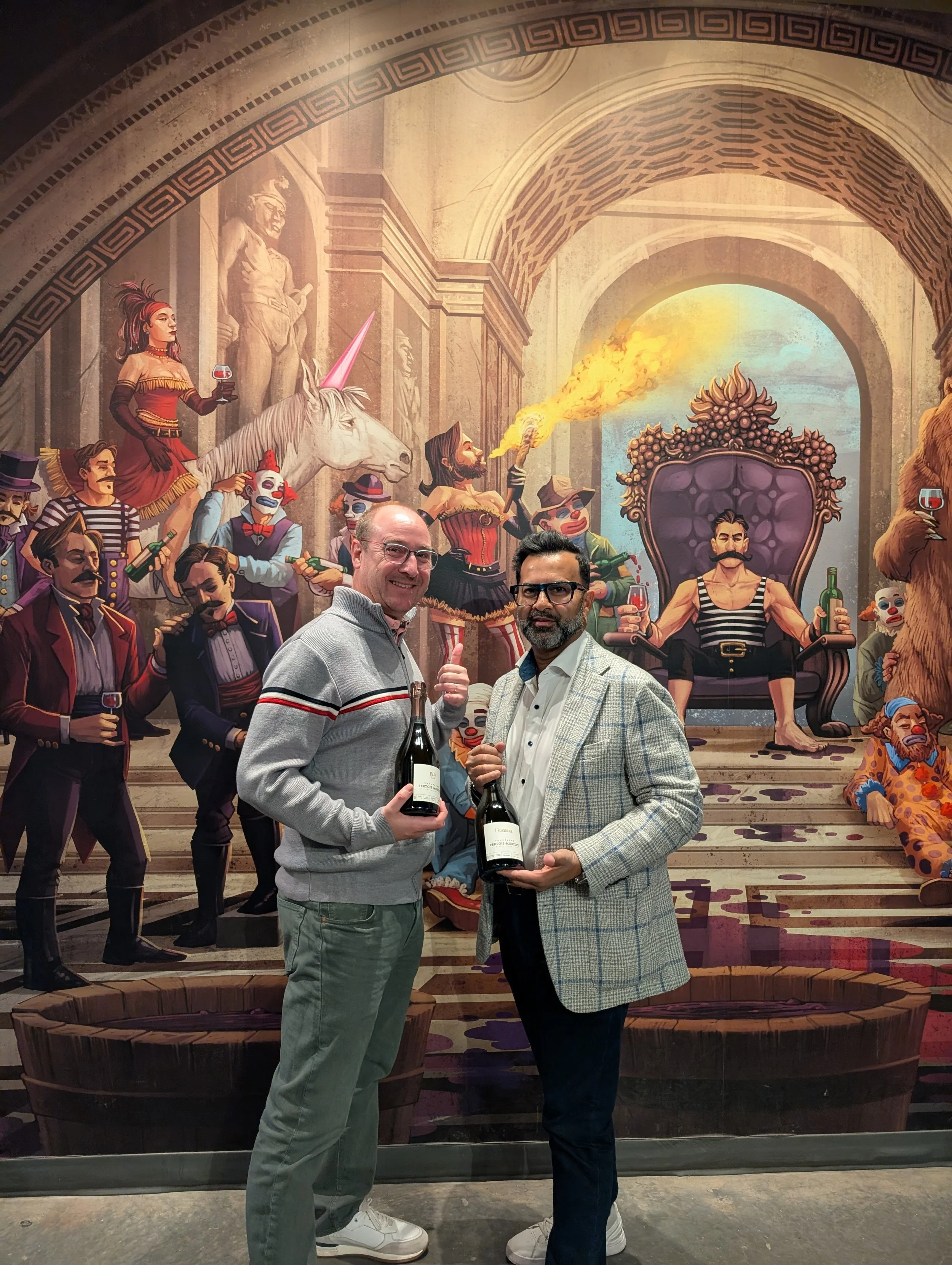 Two men standing in front of a colorful mural, holding wine bottles and giving thumbs up. The mural depicts a circus scene with clowns, a unicorn, a ringmaster, and a man on a throne.