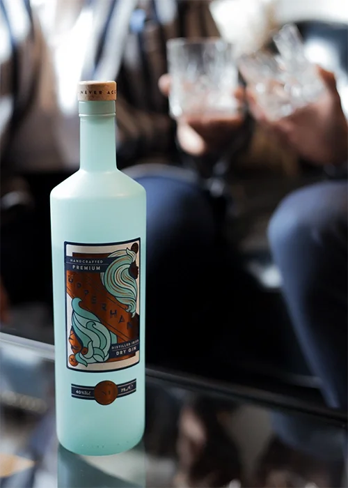 A bottle of handcrafted premium dry gin with a label featuring a stylized mermaid and ocean waves, on a table with people holding glasses in the background.