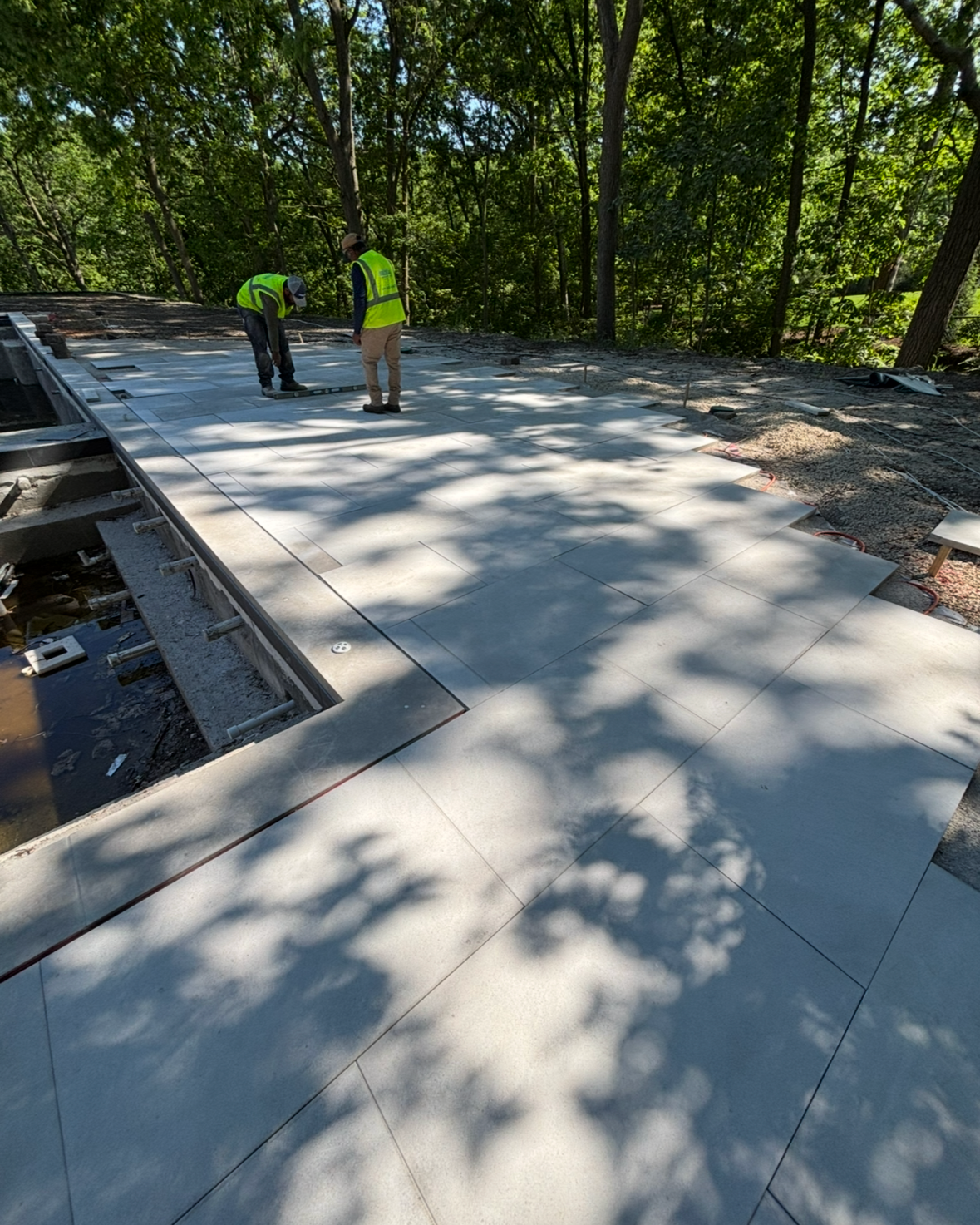 Two construction workers in safety vests and helmets installing large tiles on a roof surrounded by trees.