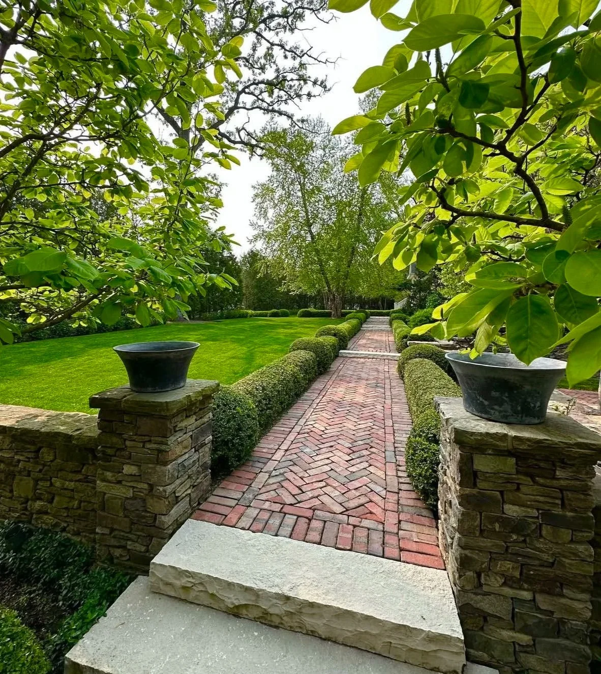 A brick pathway leading through a well-manicured garden with lush green grass, trimmed bushes, and large trees, surrounded by stone and brick accents and black planters on stone pedestals.