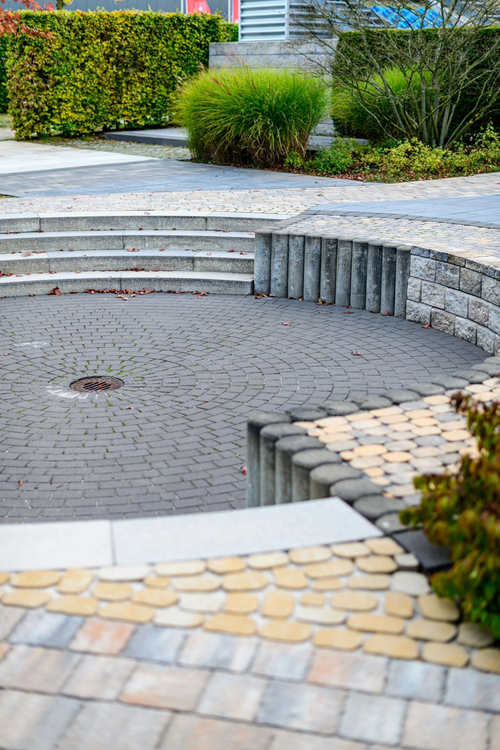 Stone and brick outdoor courtyard with curved steps, circular patterned paving, and lush green bushes and plants.