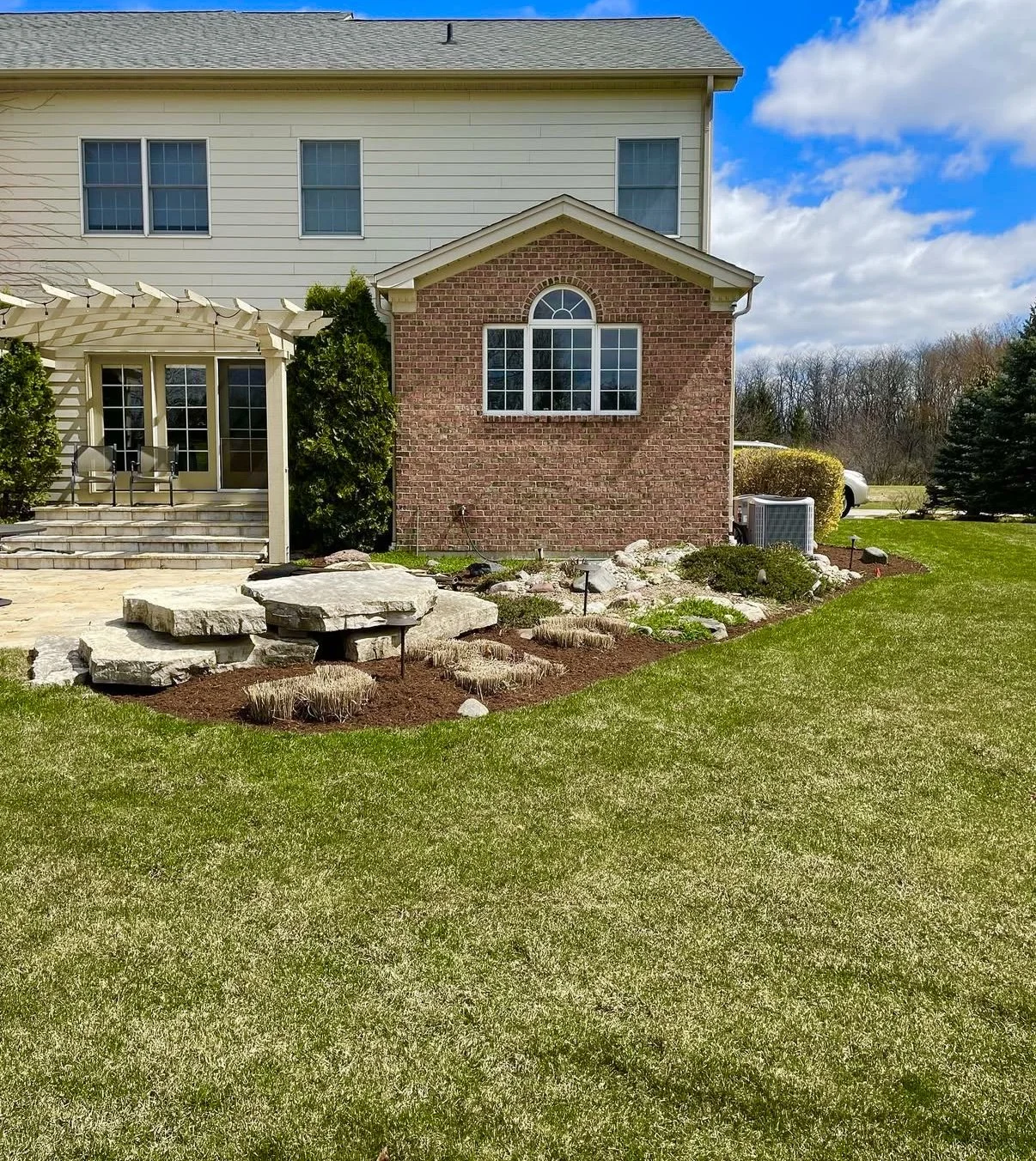 Backyard with a lawn, stone patio, garden bed with mulch and rocks, brick and siding house, and blue sky.