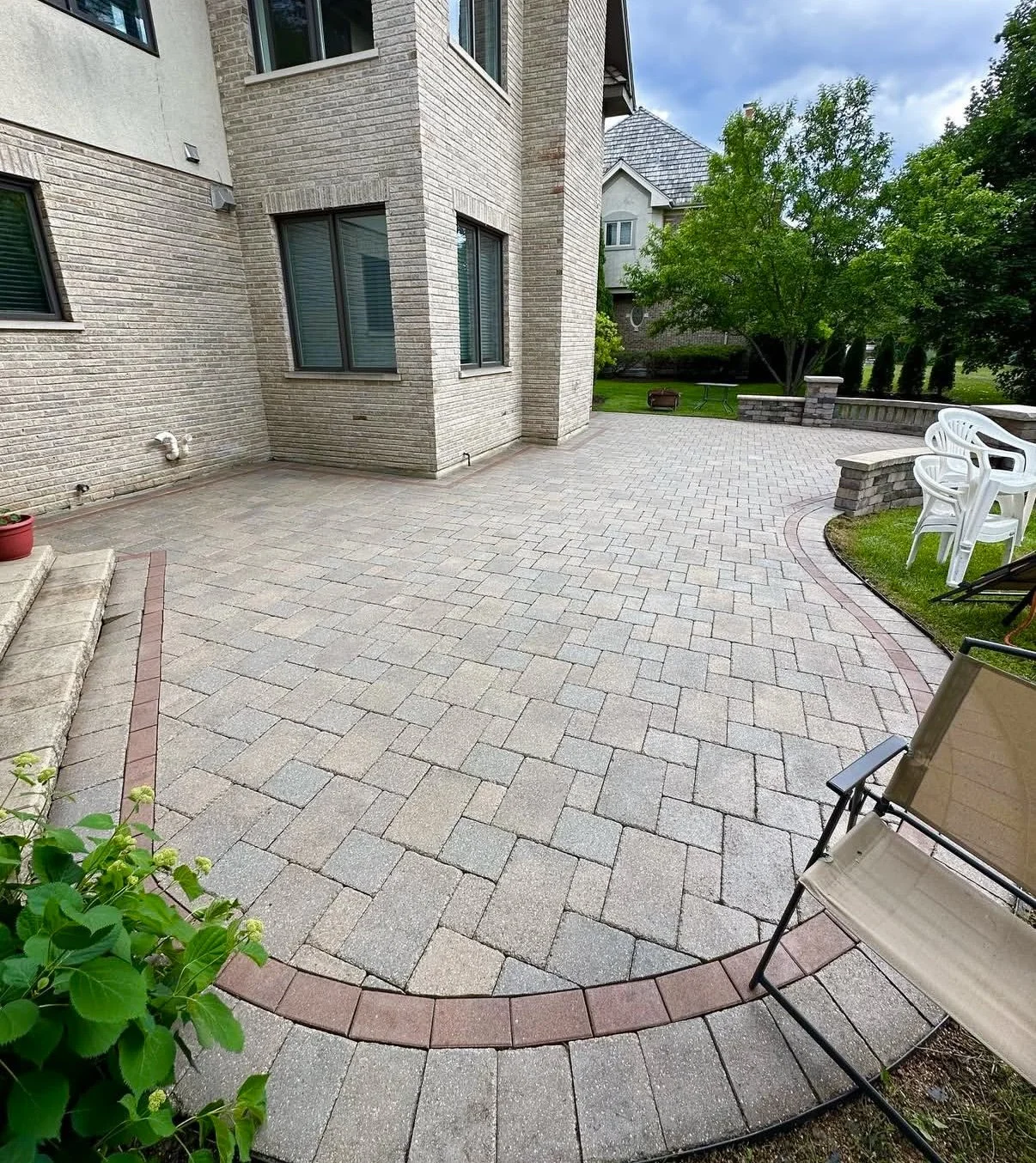 Backyard patio with brick pavers, a curved red brick border, and outdoor furniture including white plastic chairs and a beige folding chair near a brick house with large windows, a small set of stairs, a green lawn, trees, and a partly cloudy sky.