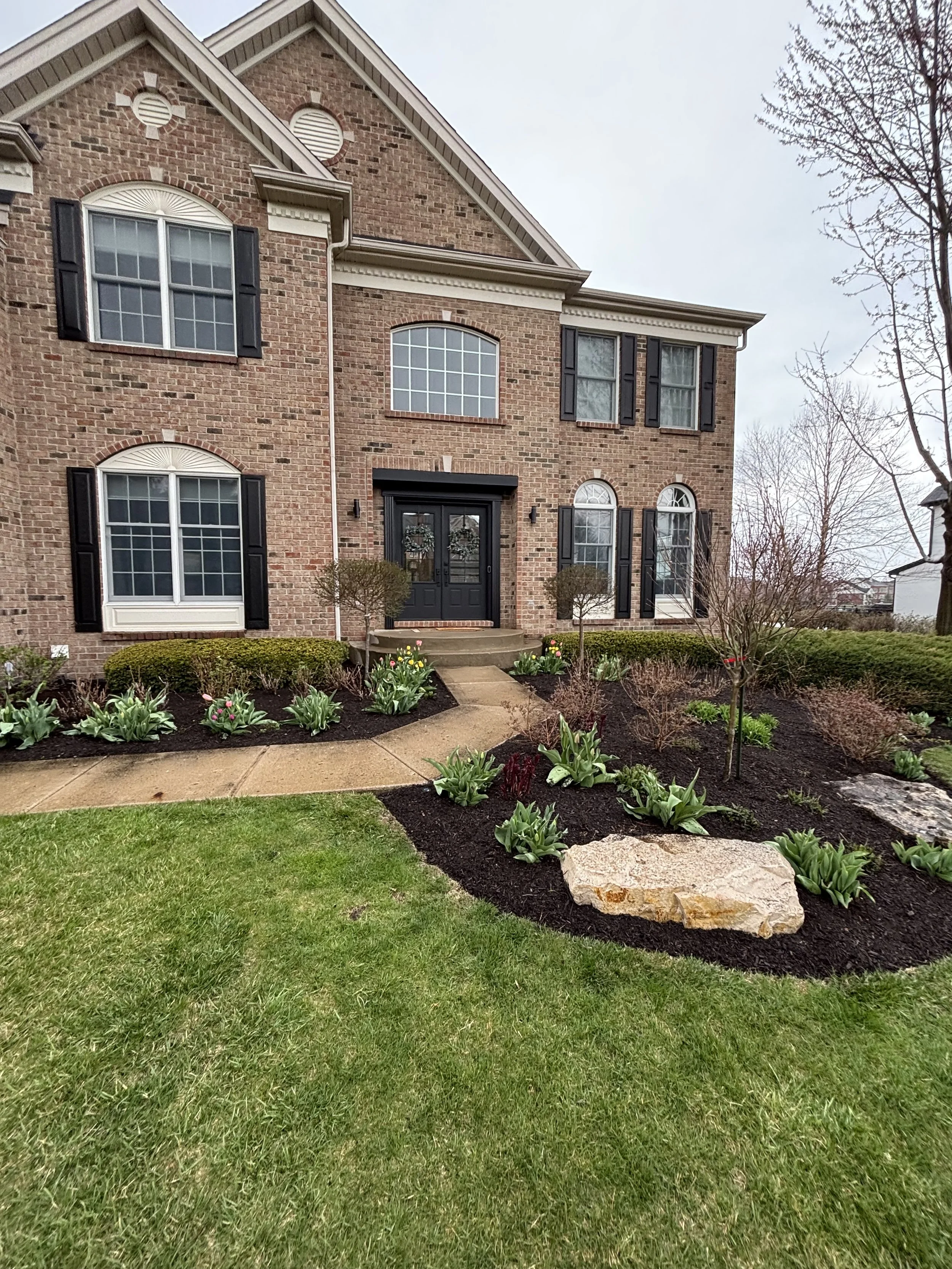 Front view of a two-story brick house with black shutters, black front door, landscaped garden with plants, shrubs, flowers, a large stone, and a concrete walkway leading to the entrance.