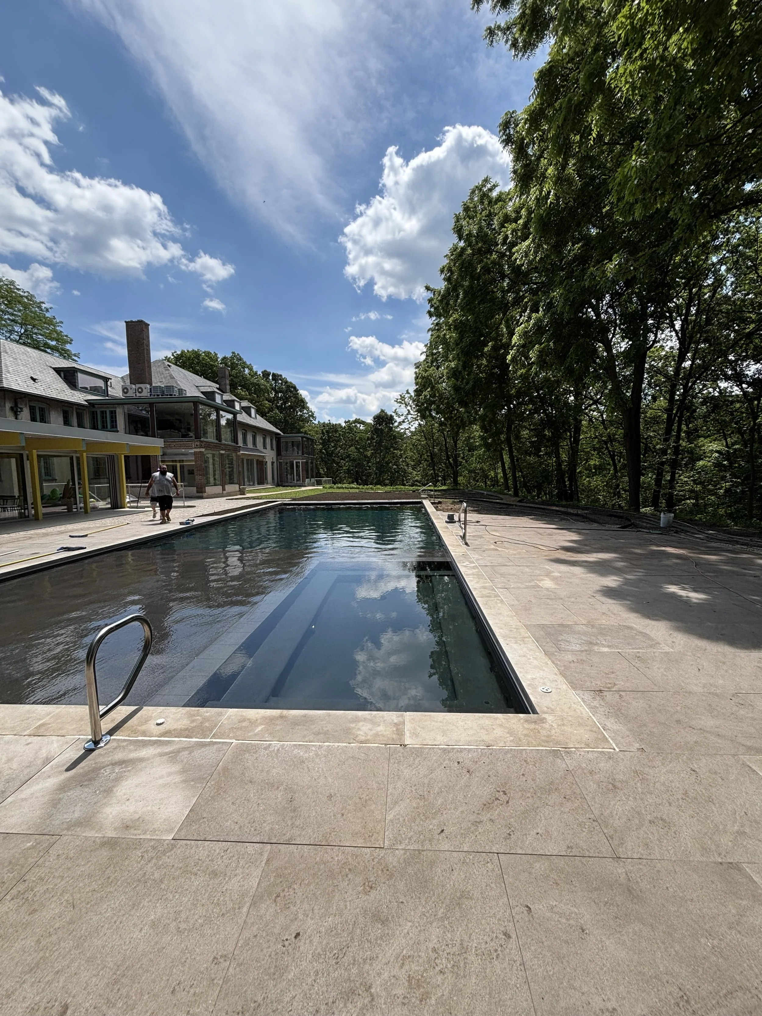 Empty swimming pool next to a modern house with a backyard patio, surrounded by trees and a partly cloudy sky.
