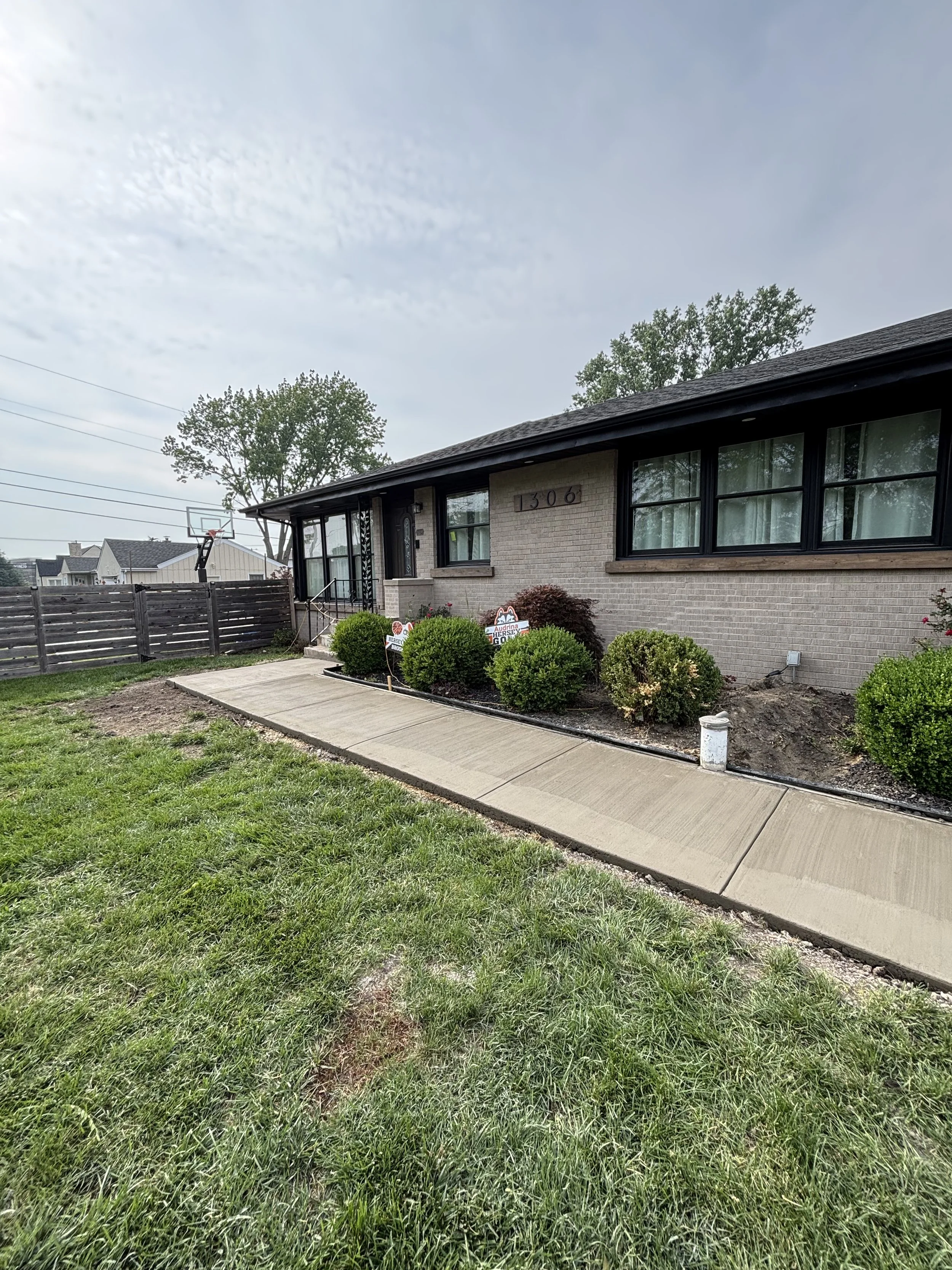 Front view of a single-story brick house with a concrete sidewalk walkway, shrubs, and a fenced backyard with a basketball hoop.