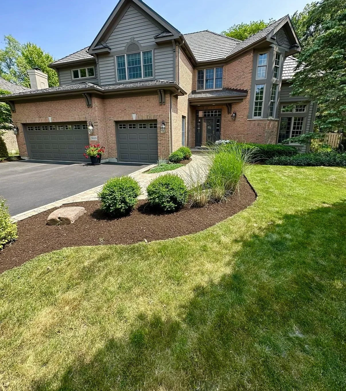 Front view of a two-story house with brick and gray siding, a paved driveway, well-maintained lawn, and landscaped garden with shrubs and plants.