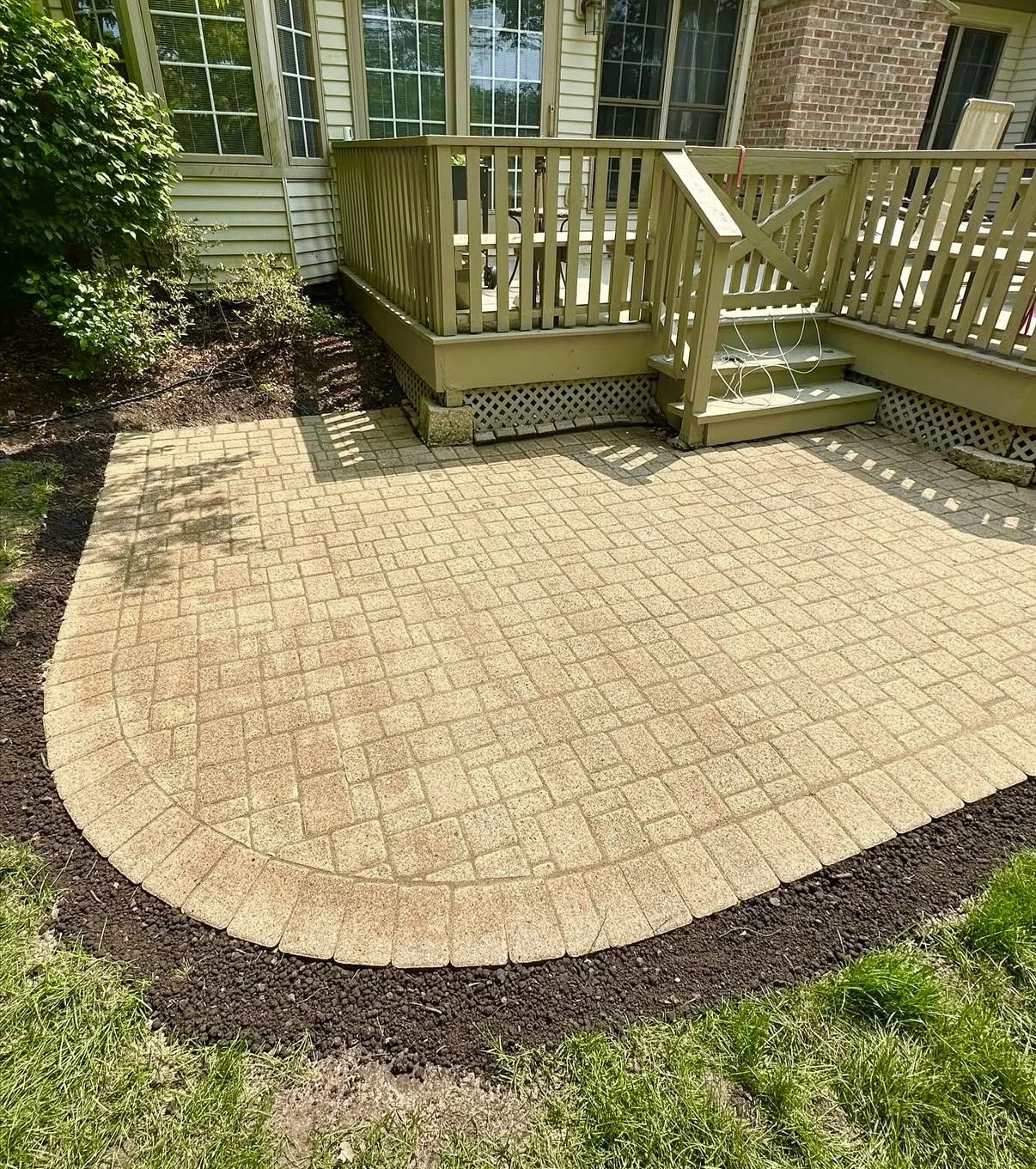 Backyard patio with brick pavers, surrounded by a garden bed with soil and grass, adjacent to a house with a wooden deck and railing, and stairs leading down to the patio.