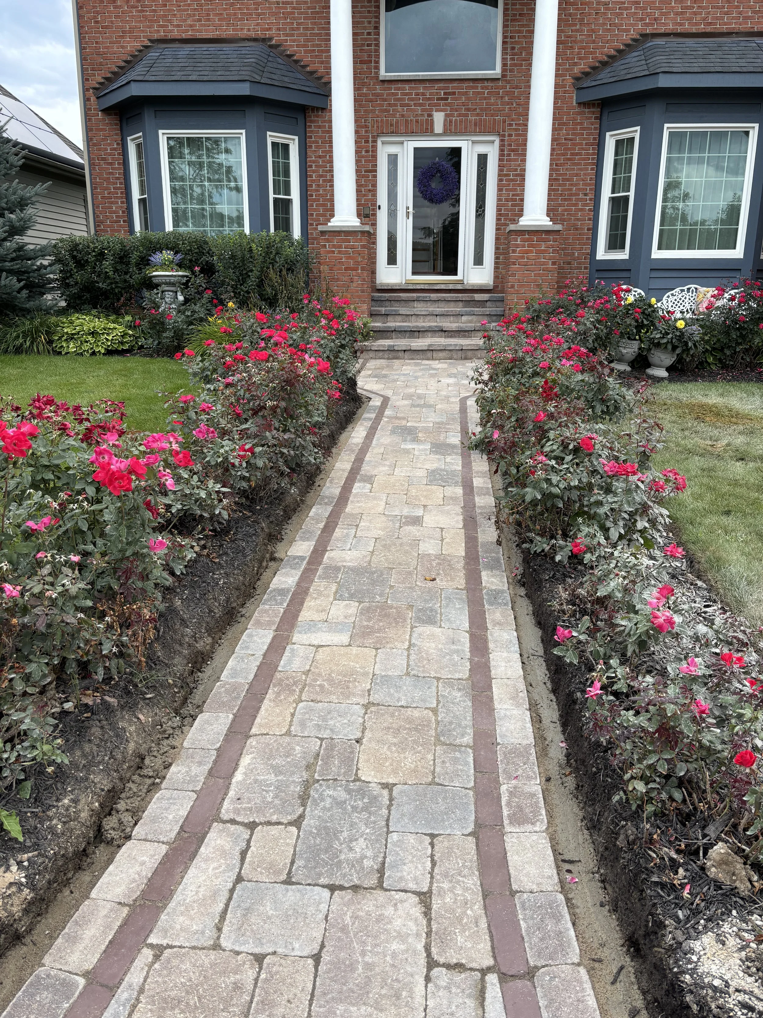 Brick walkway leading to the front door of a house, flanked by flower beds with pink flowering plants and edged with bricks.