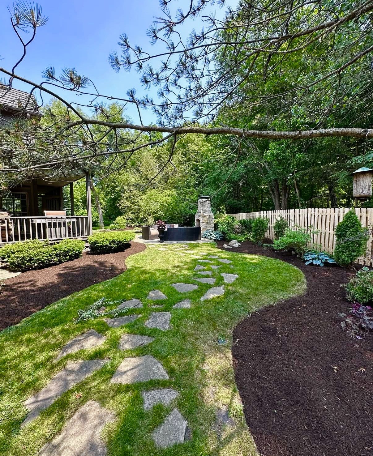 A landscaped backyard with a stone pathway, green grass, shrubs, and trees, with a porch on the left side and a stone fireplace at the back, enclosed by a wooden fence.