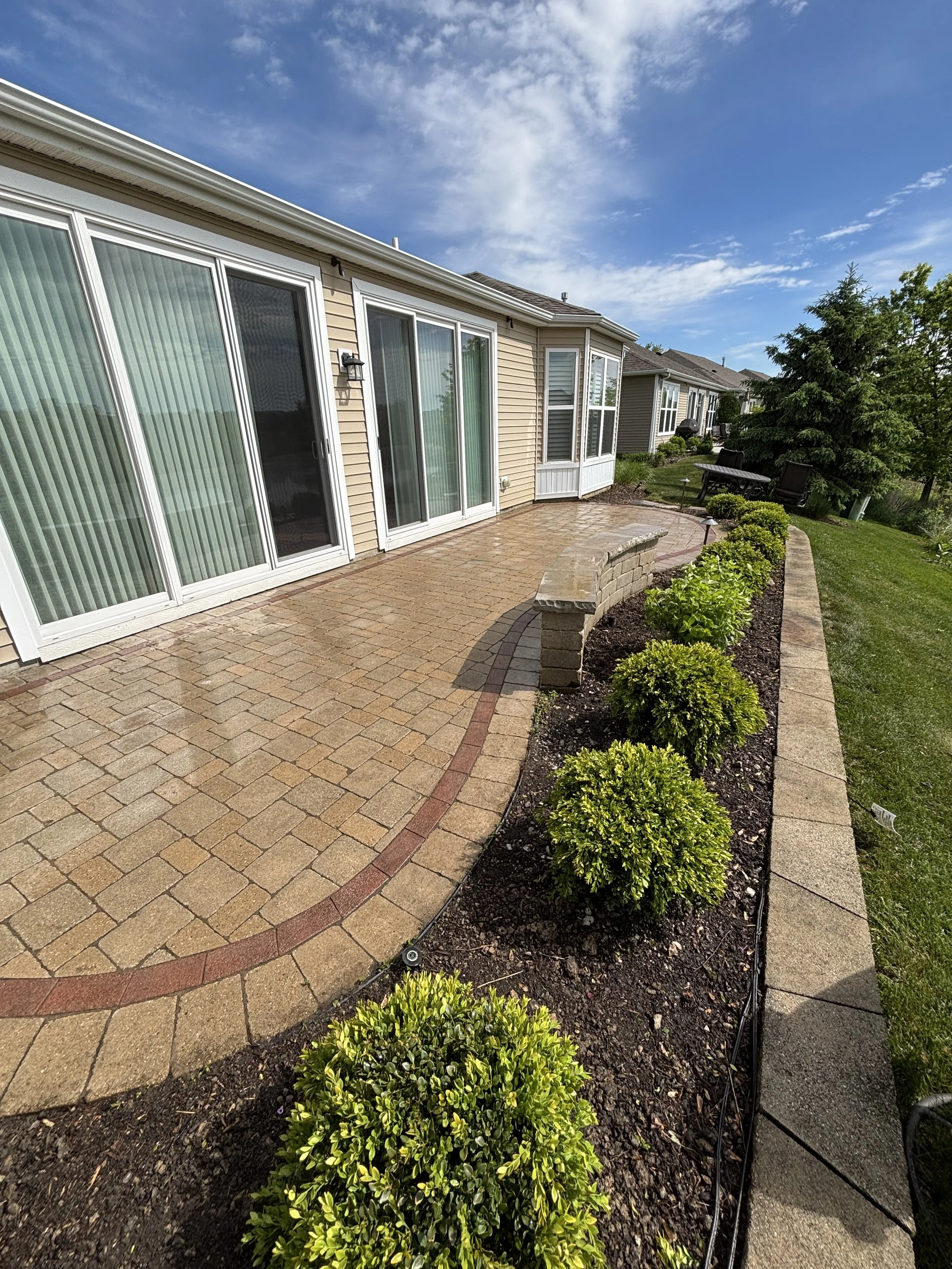 Backyard patio with stone pavers, flower bed with green shrubbery, sliding glass doors, and outdoor seating under trees on a sunny day.