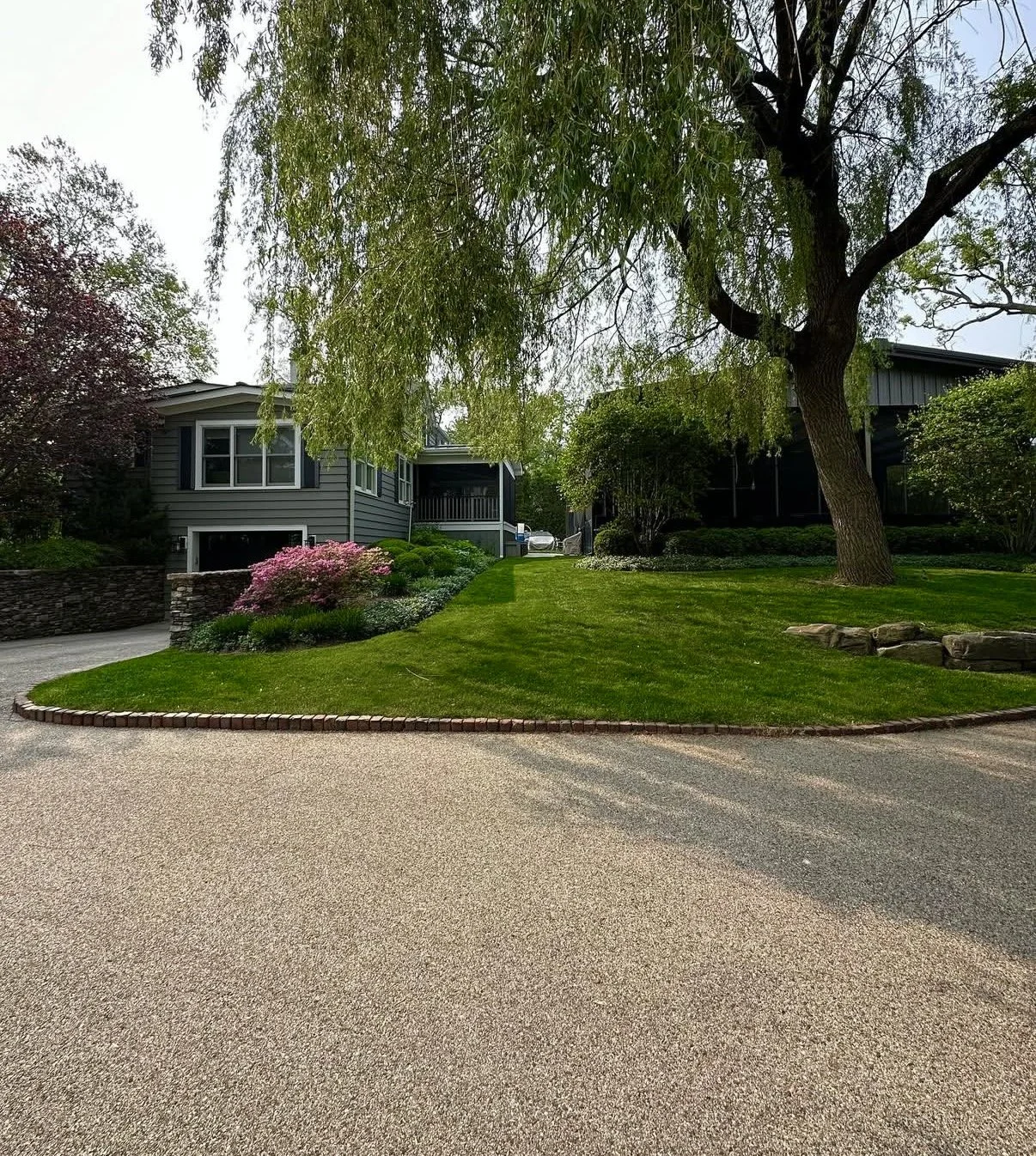 A landscaped front yard with green grass, a large tree, and flower bushes in front of a modern dark gray house with multiple windows and a porch. The driveway curves around the lawn.