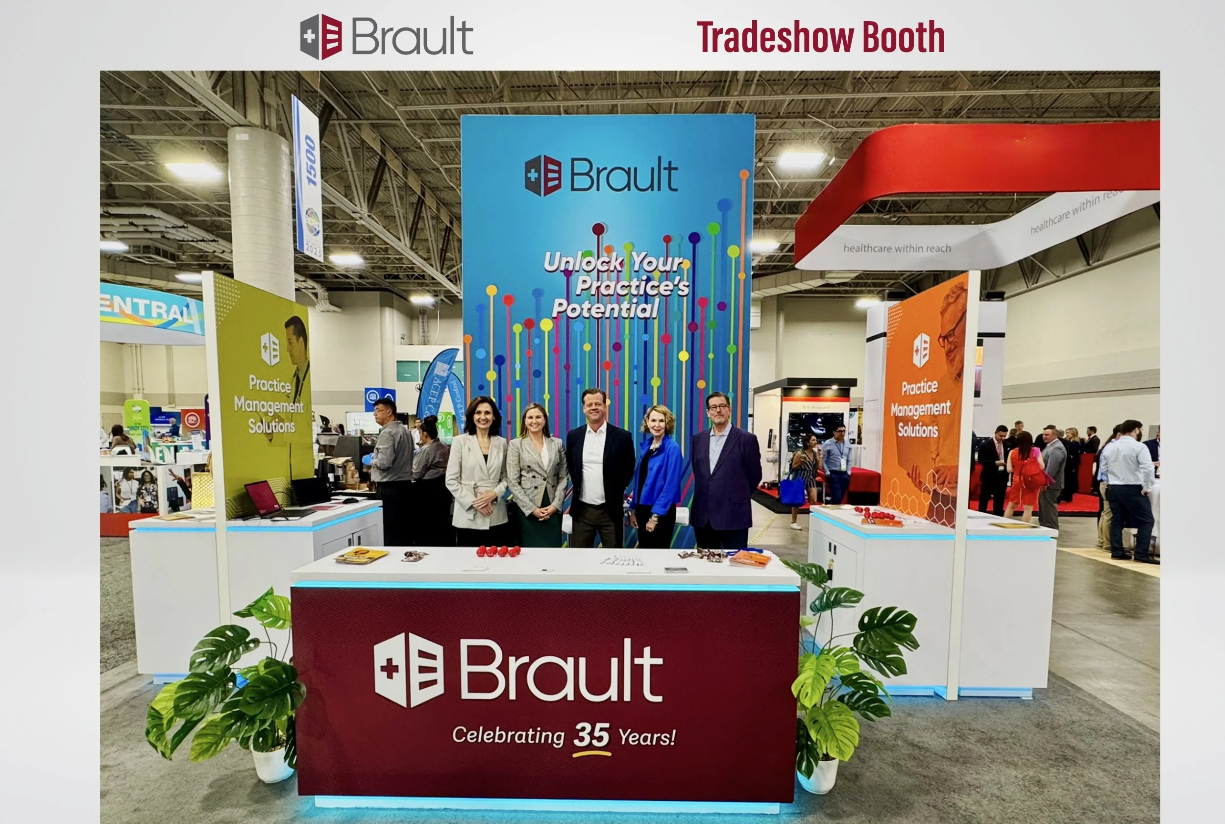 Brault booth at a trade show with a large blue background displaying the company logo and the slogan 'Unlock Your Practice's Potential.' The booth has a red front panel with the Brault logo and 'Celebrating 35 Years!' written on it. Four people stand behind the booth, smiling, with two women and two men dressed professionally. The booth includes signs for Practice Management Solutions and has various promotional materials on the tables. The trade show is busy with visitors and other booths visible in the background.