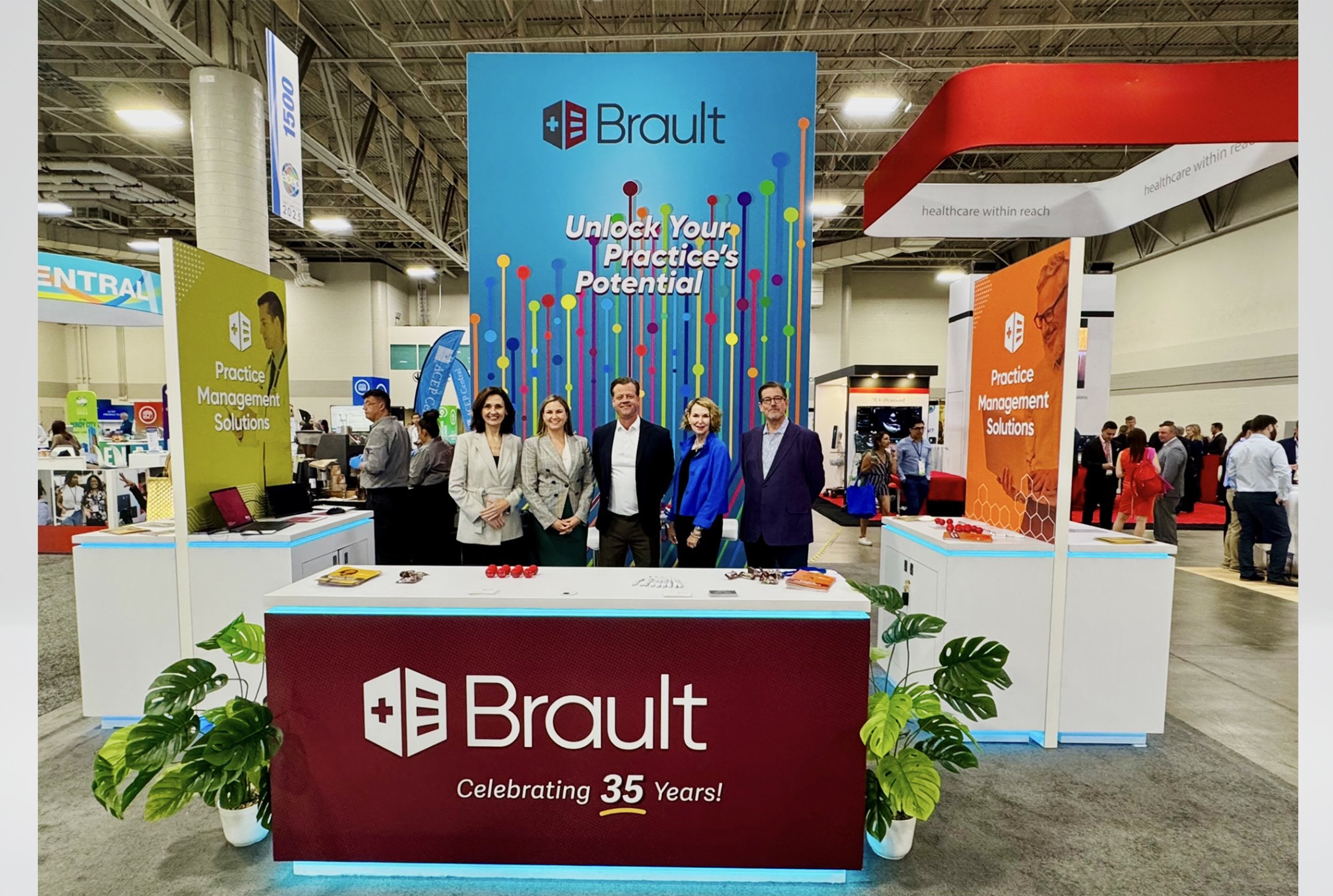 Brault booth at a trade show with a large blue background displaying the company logo and the slogan 'Unlock Your Practice's Potential.' The booth has a red front panel with the Brault logo and 'Celebrating 35 Years!' written on it. Four people stand behind the booth, smiling, with two women and two men dressed professionally. The booth includes signs for Practice Management Solutions and has various promotional materials on the tables. The trade show is busy with visitors and other booths visible in the background.