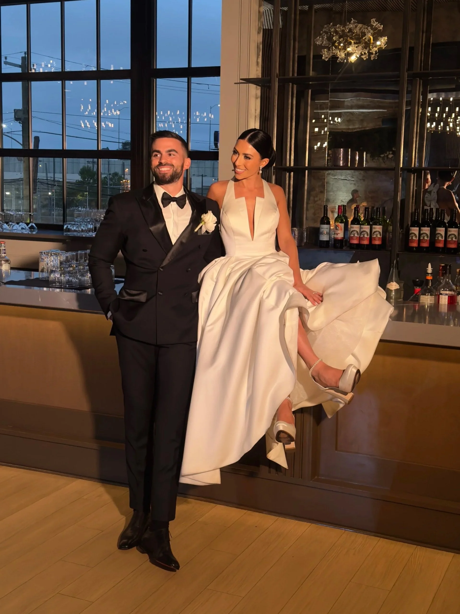 A bride and groom pose together in a stylish indoor setting, with the bride sitting on a bar counter and the groom standing beside her, both dressed in formal wedding attire.