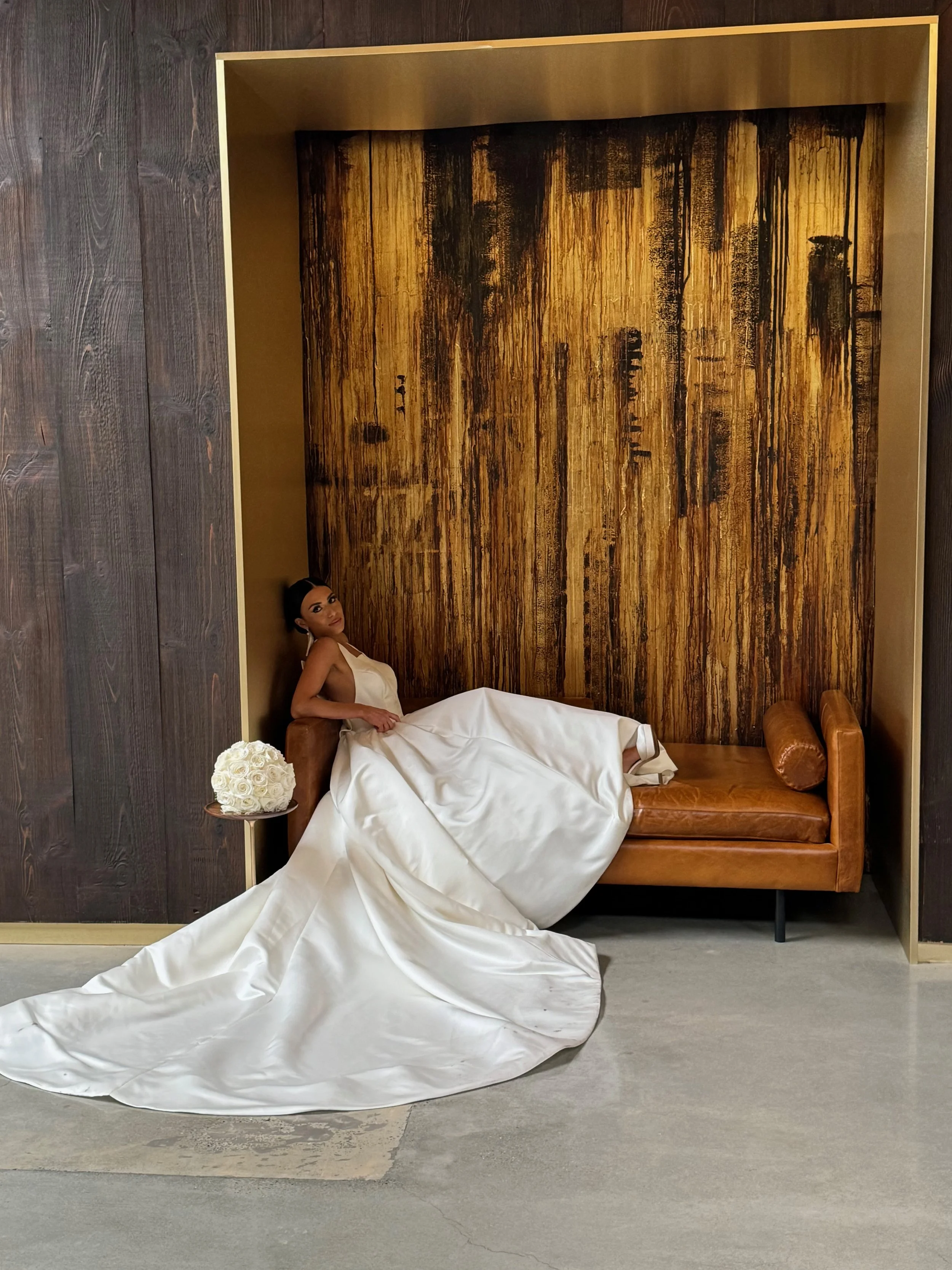 A woman in a white dress sits on a brown leather bench inside a decorative wooden alcove, with a bouquet of white roses on a small table beside her.