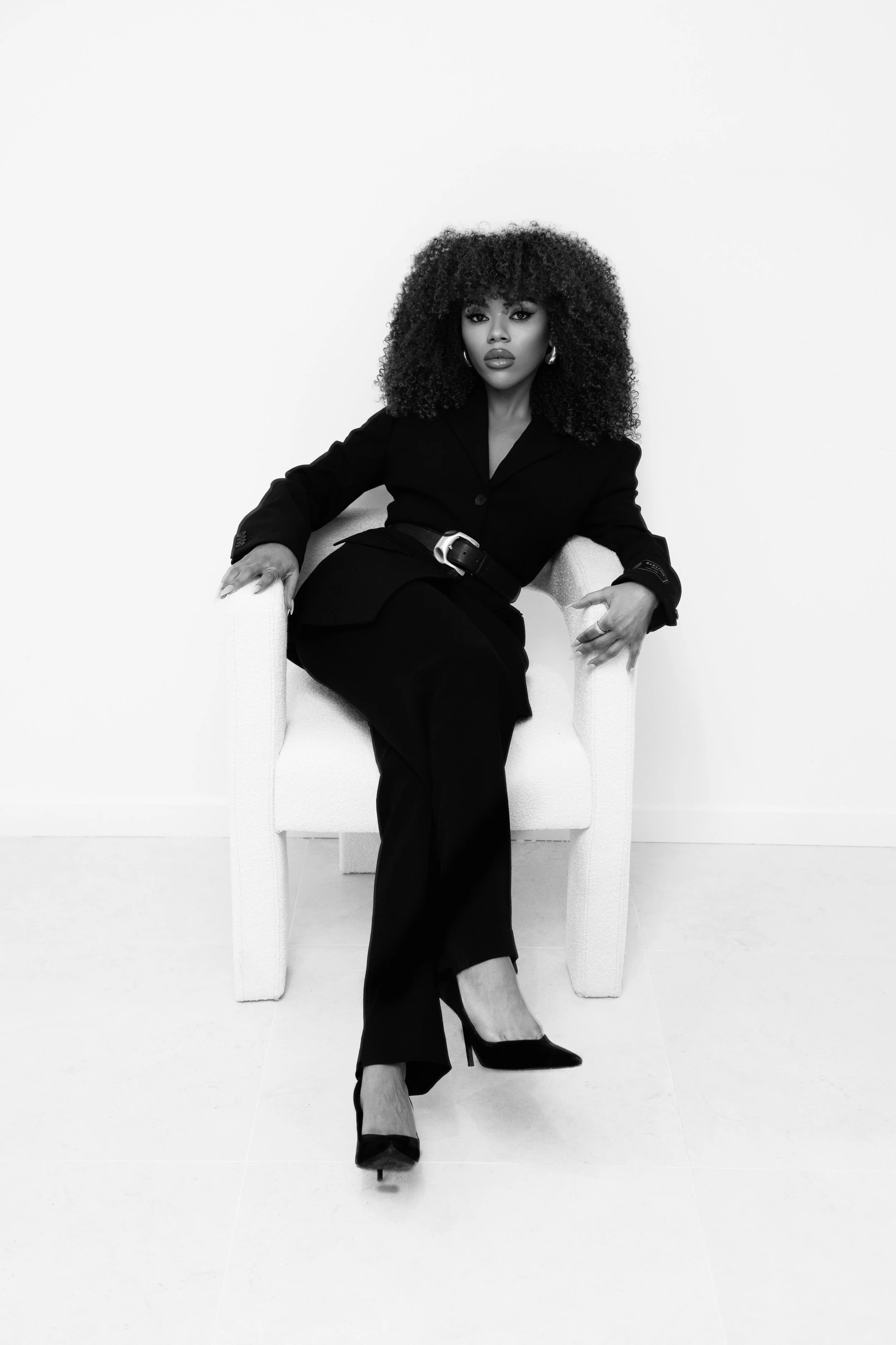 A woman with curly hair wearing a black blazer, belt, and pants, sitting confidently on a white chair against a plain white background