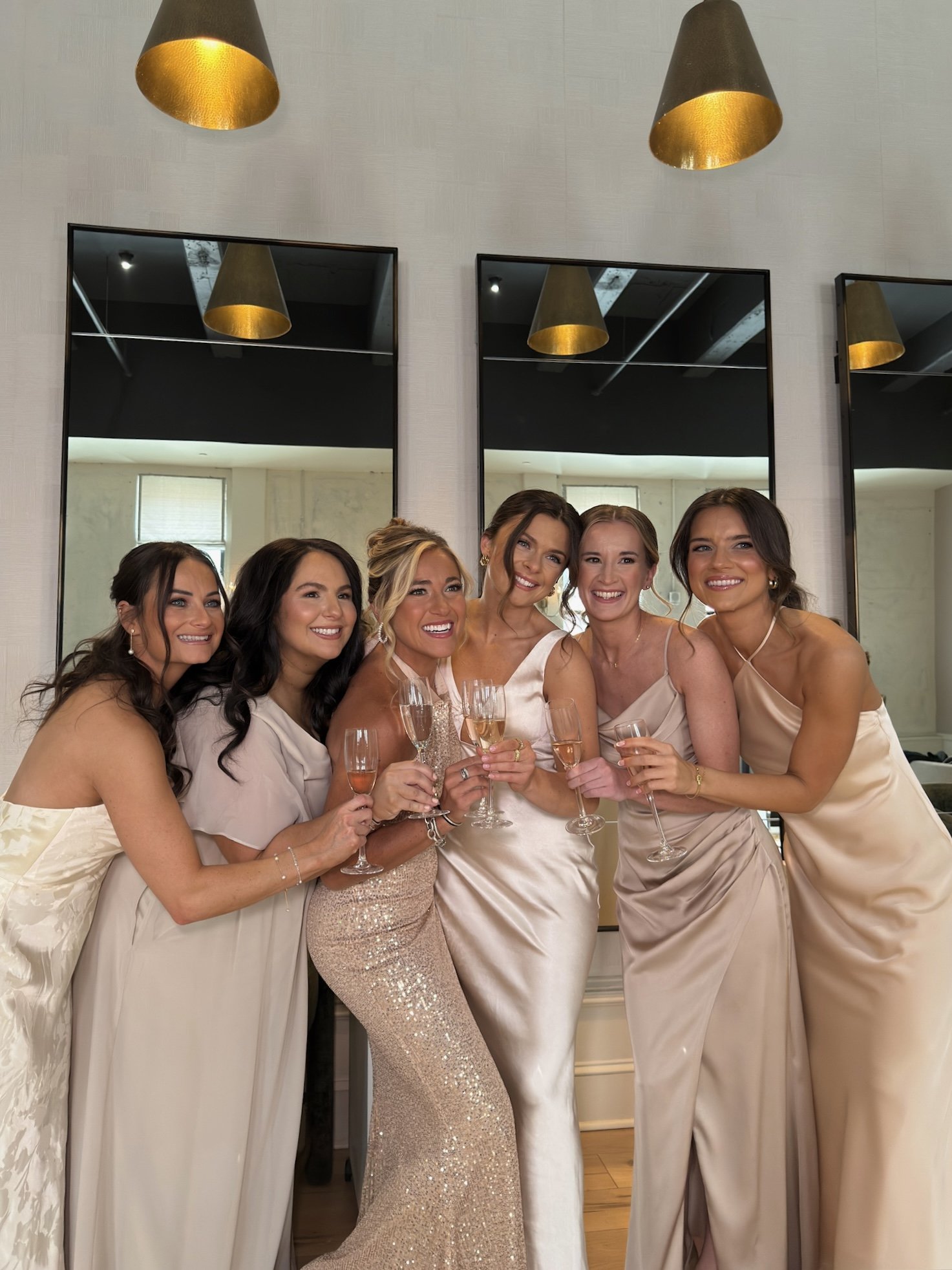 Six women in elegant dresses holding champagne glasses, smiling at a celebration or wedding reception, standing in front of mirrors on a white wall.