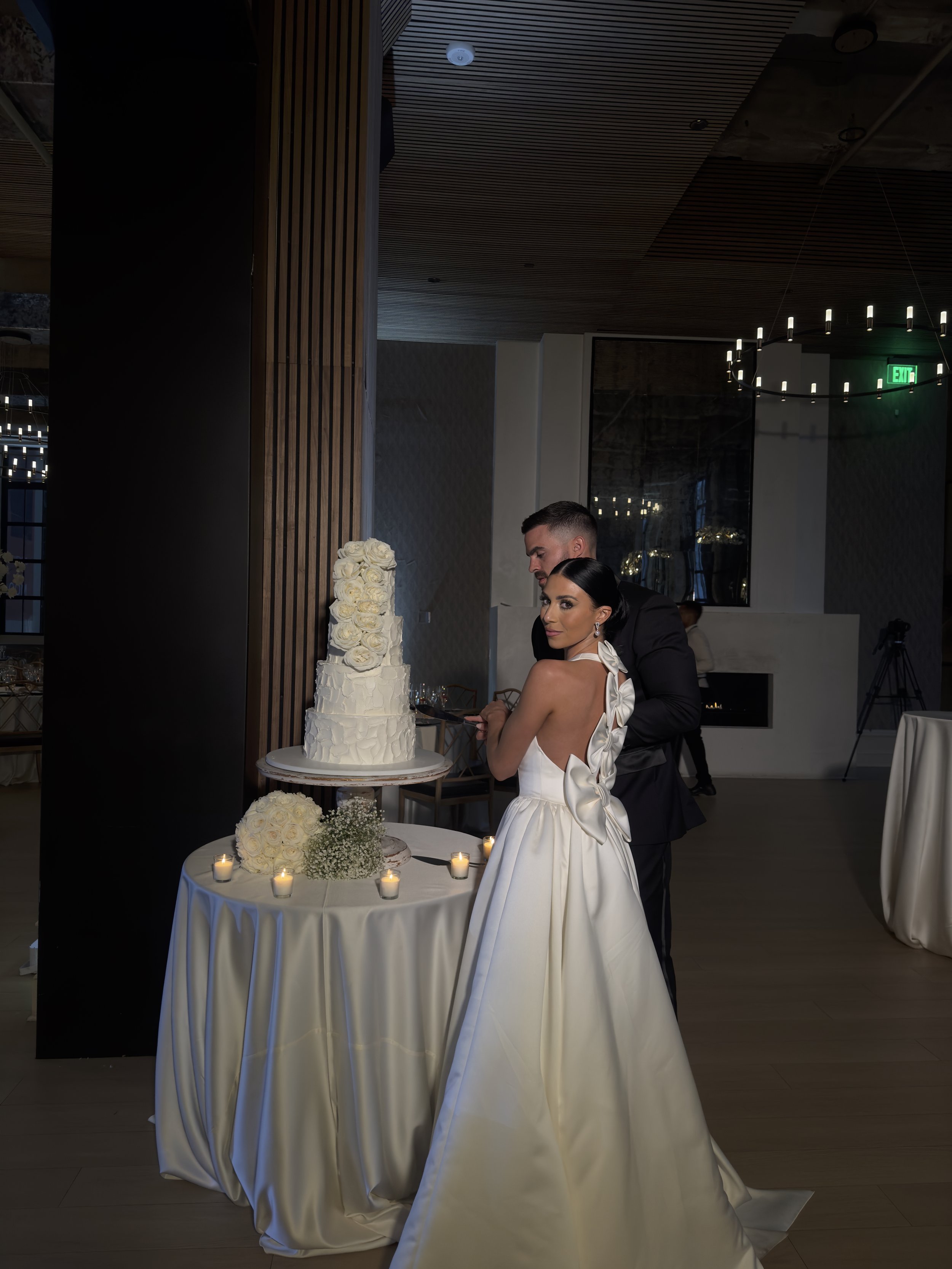 Bride and groom cutting a wedding cake at reception. The bride is in a white gown with a large bow at the back, and the groom is in a black tuxedo. The wedding cake is tall with white roses, placed on a table with candles and flowers.