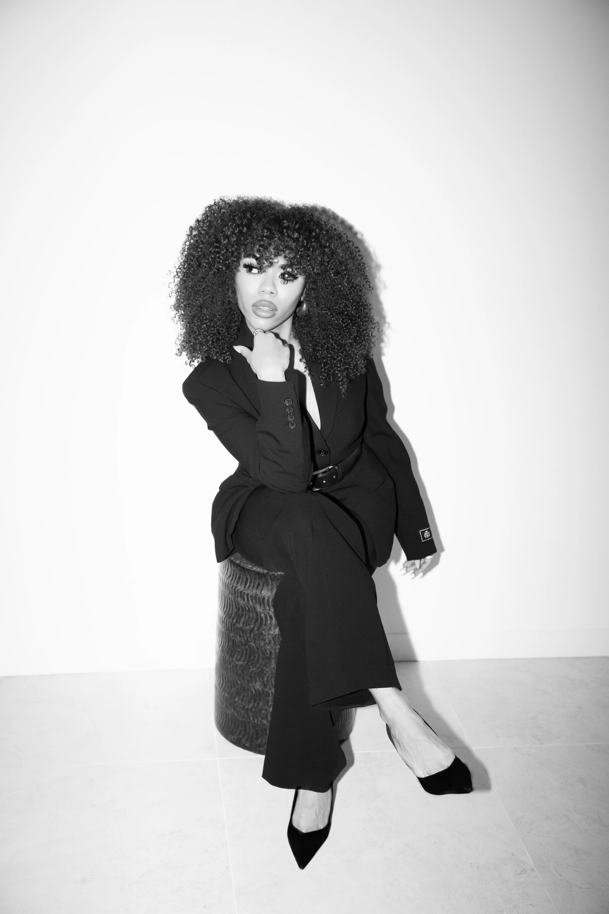 A woman with curly hair posing against a plain background. She's dressed in a dark suit, with one hand resting on her chin, and wearing high heels.