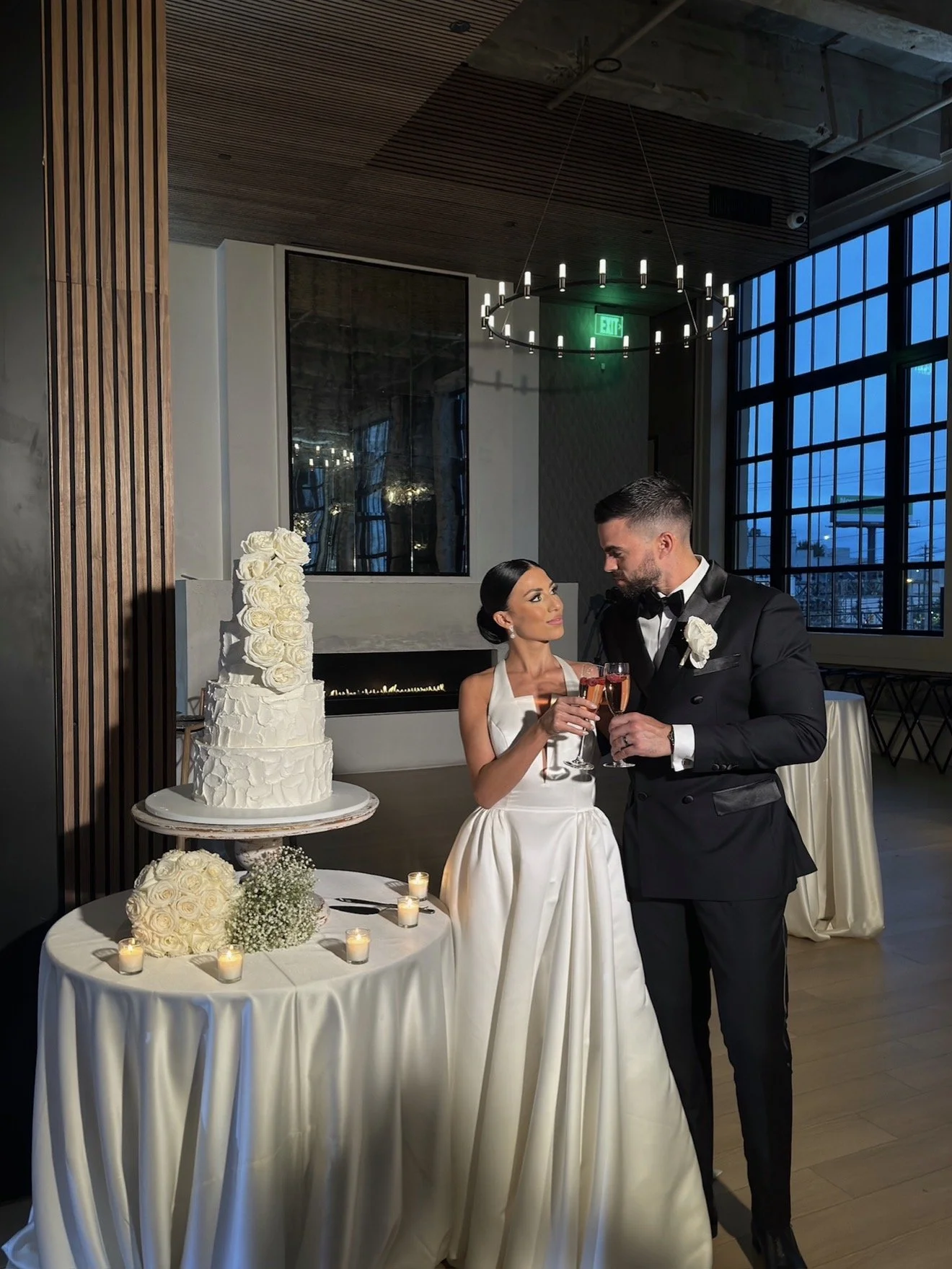 A bride and groom in wedding attire standing near a wedding cake, raising glasses in a toast at their wedding reception indoors.