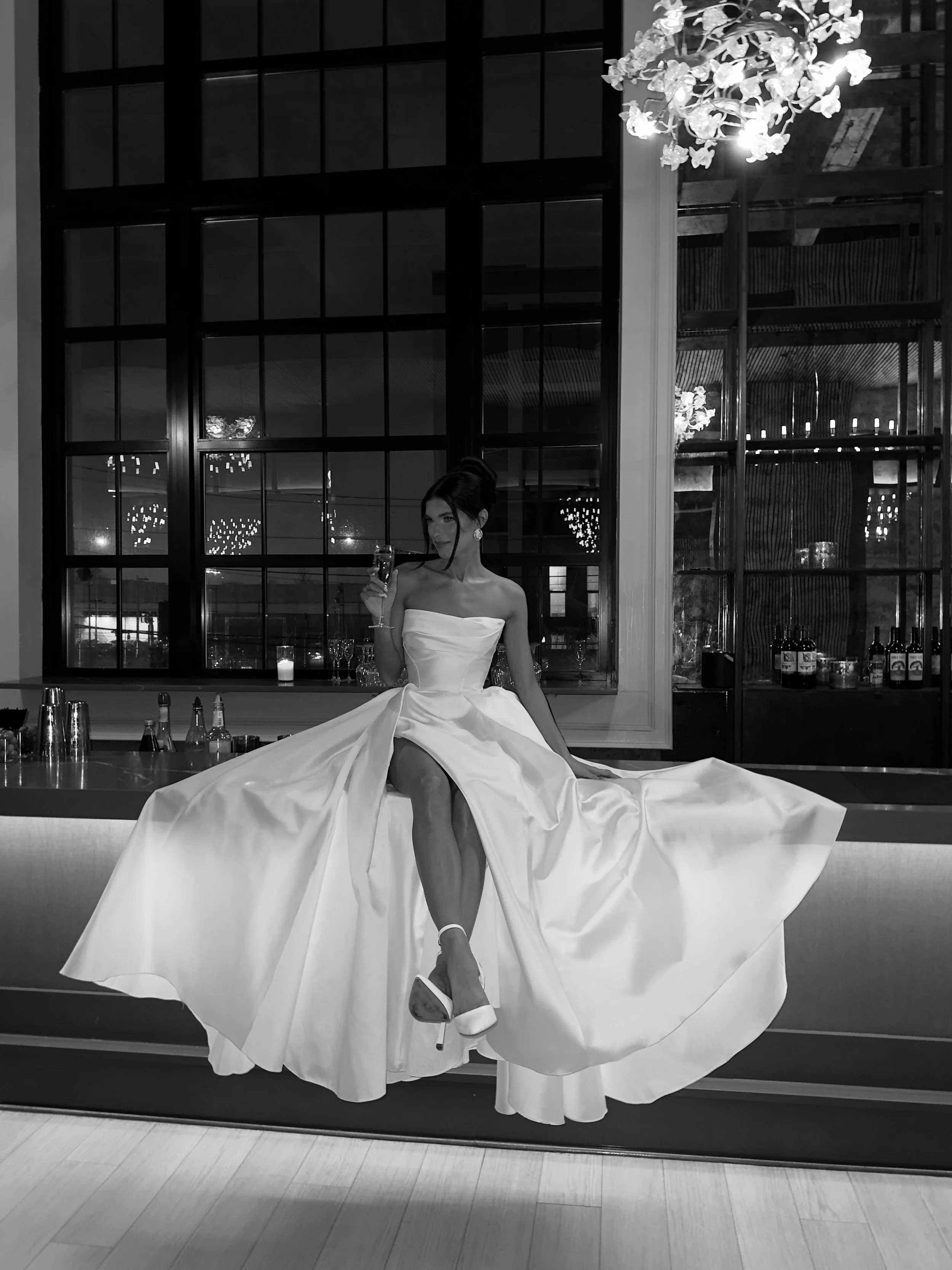 A woman in a strapless wedding dress sitting on a bar counter, sipping a drink in a stylish venue with large window panes and hanging chandeliers.
