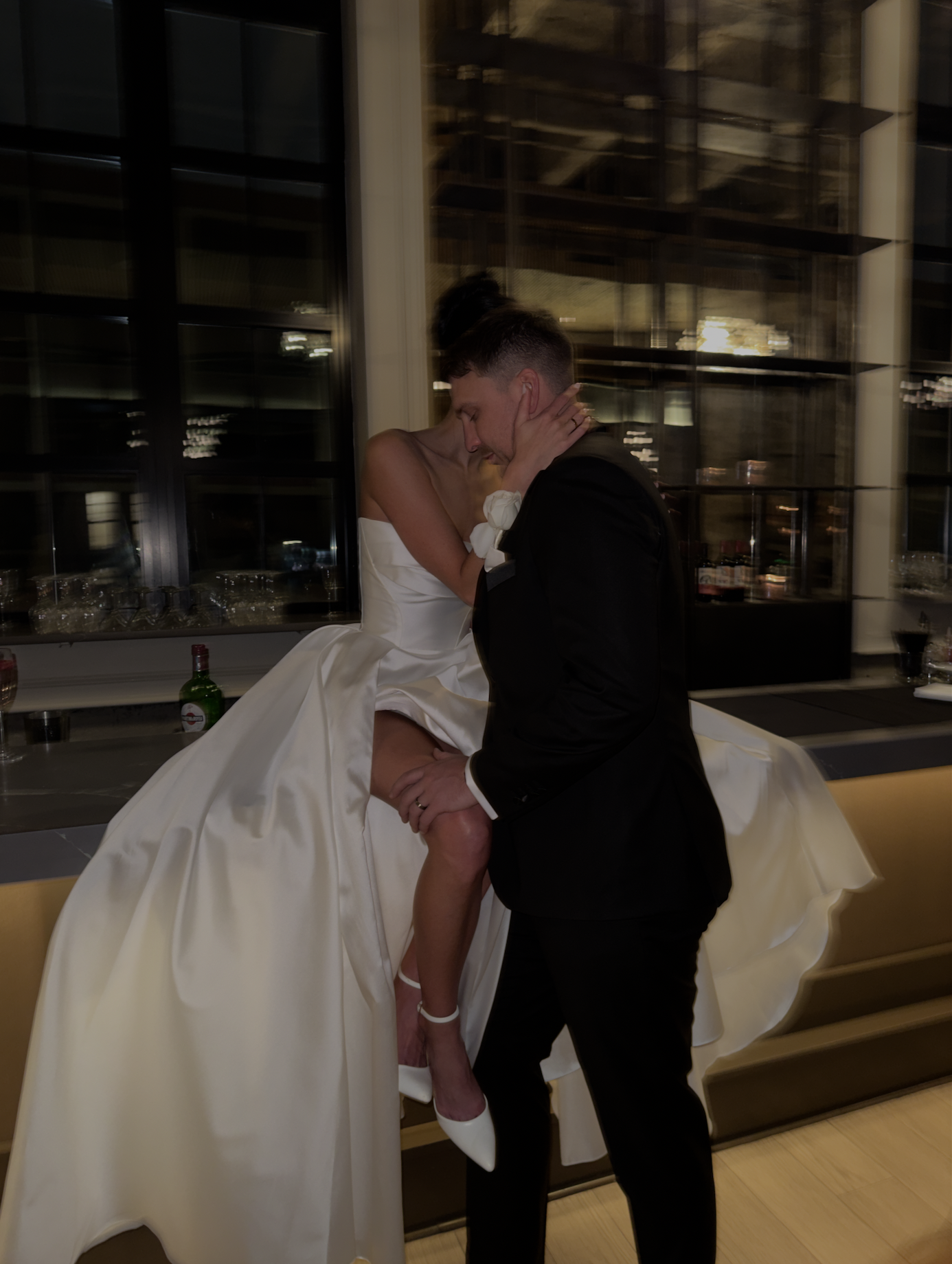 A bride and groom are kissing passionately at their wedding reception, with the bride sitting on a ledge and the groom standing close, holding her leg.