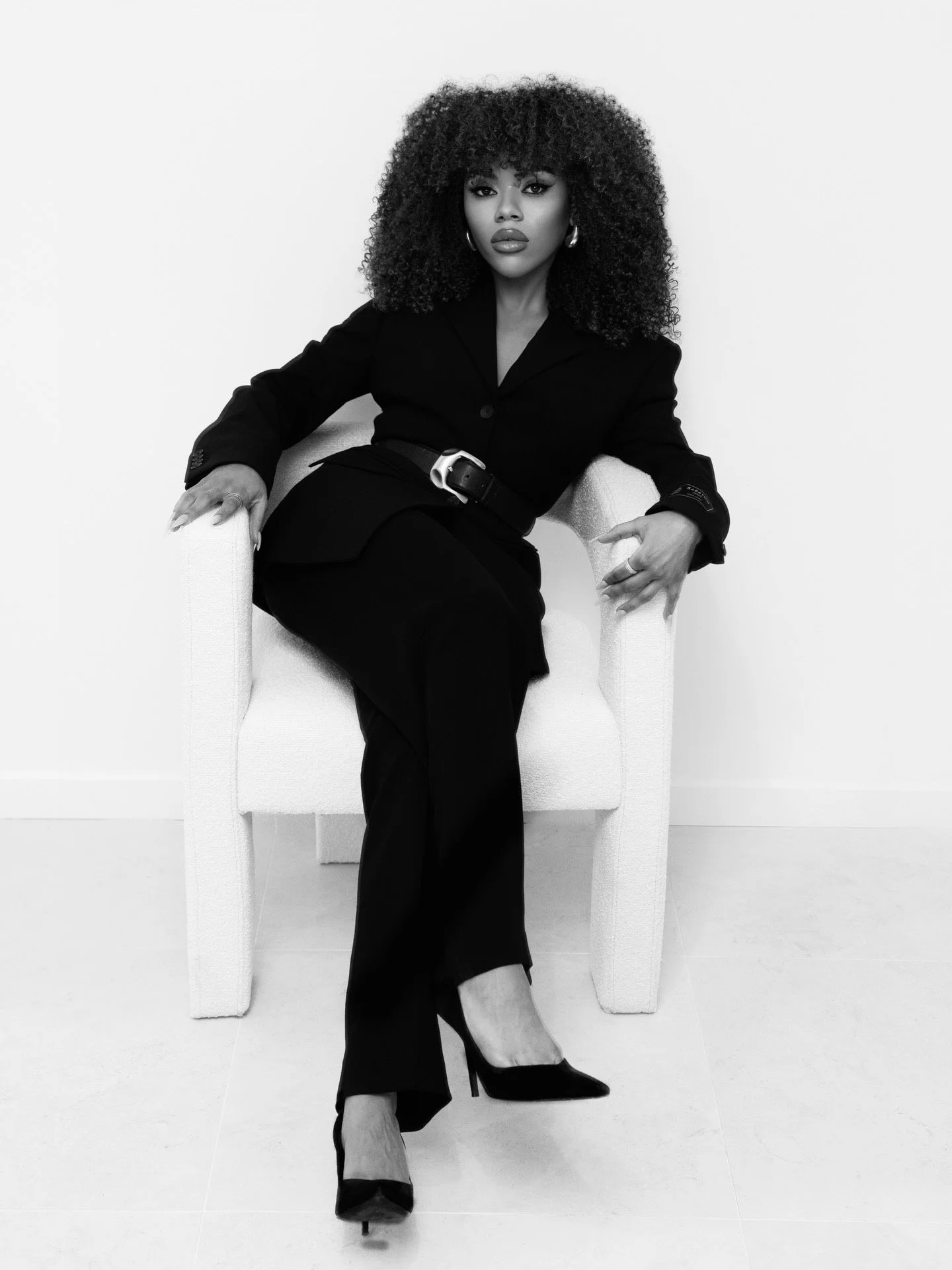 Black and white photograph of a woman sitting in a white armchair, wearing a black blazer, black pants, and high heels. She has curly hair and is looking directly at the camera.