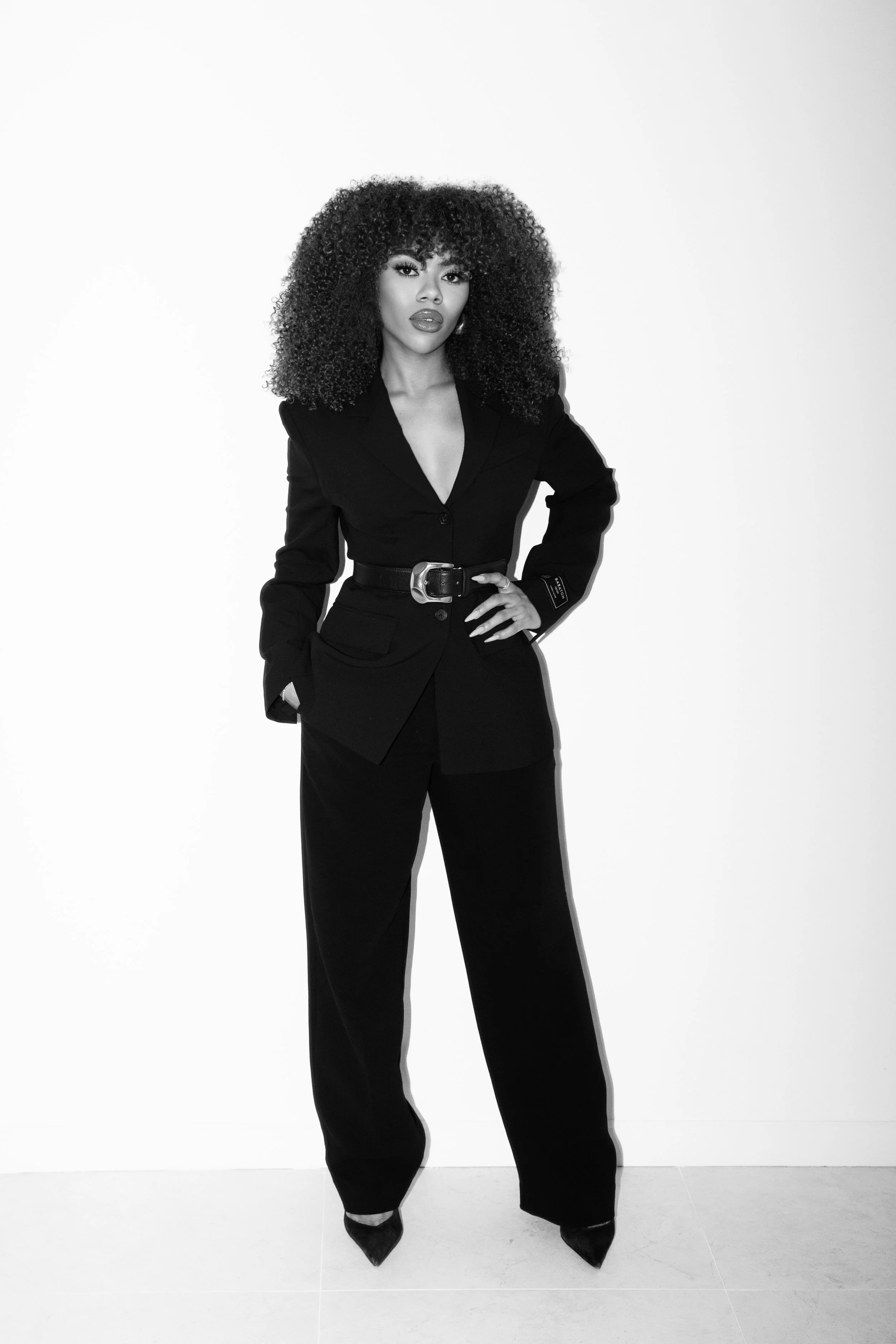 A woman in a black suit standing against a plain white background. She has curly hair and bold makeup with lipstick, posing confidently with one hand on her hip.