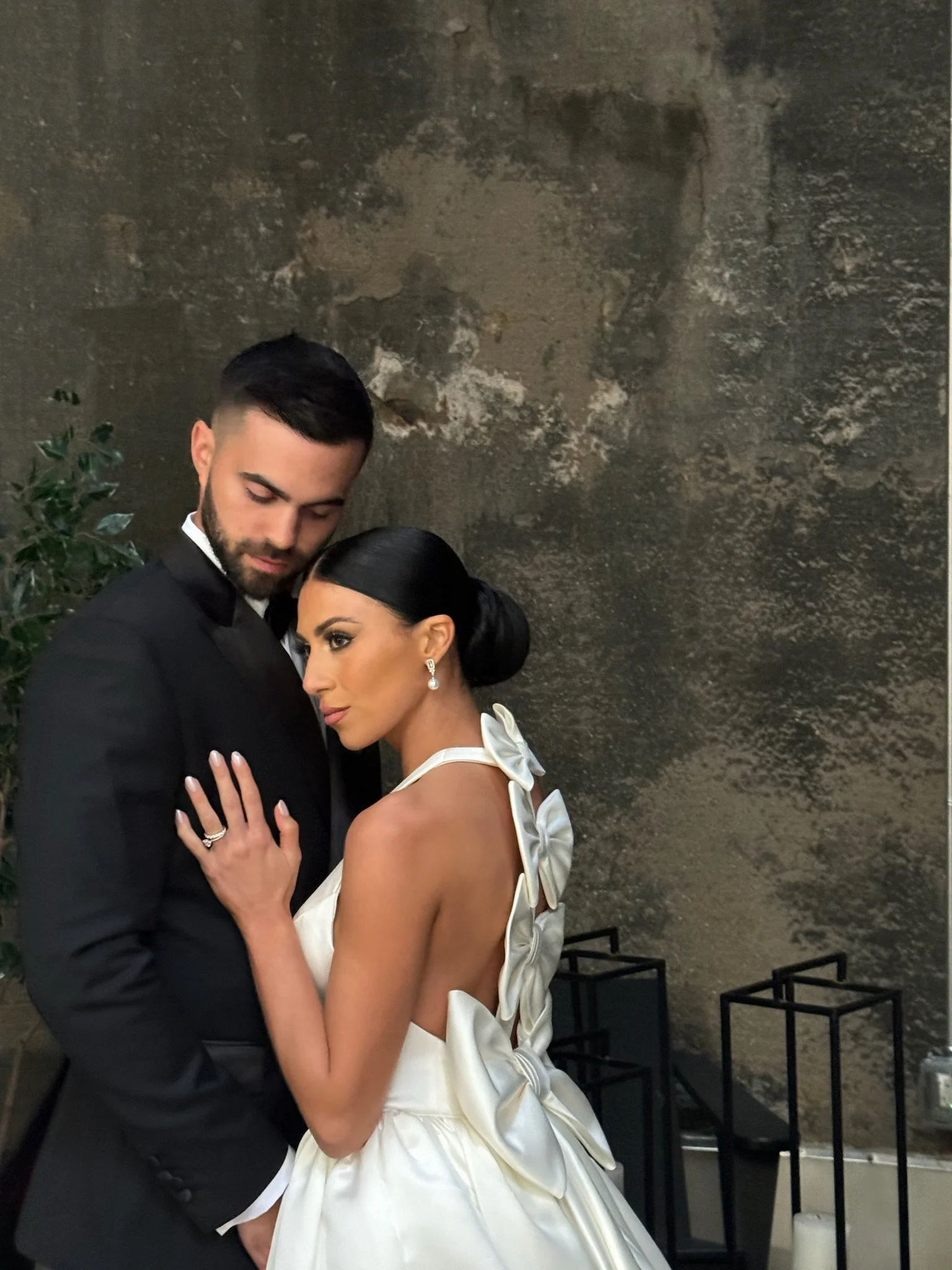 A couple dressed in formal attire, the woman in a white gown with bows on her back, and the man in a black tuxedo, standing close together against a textured wall.