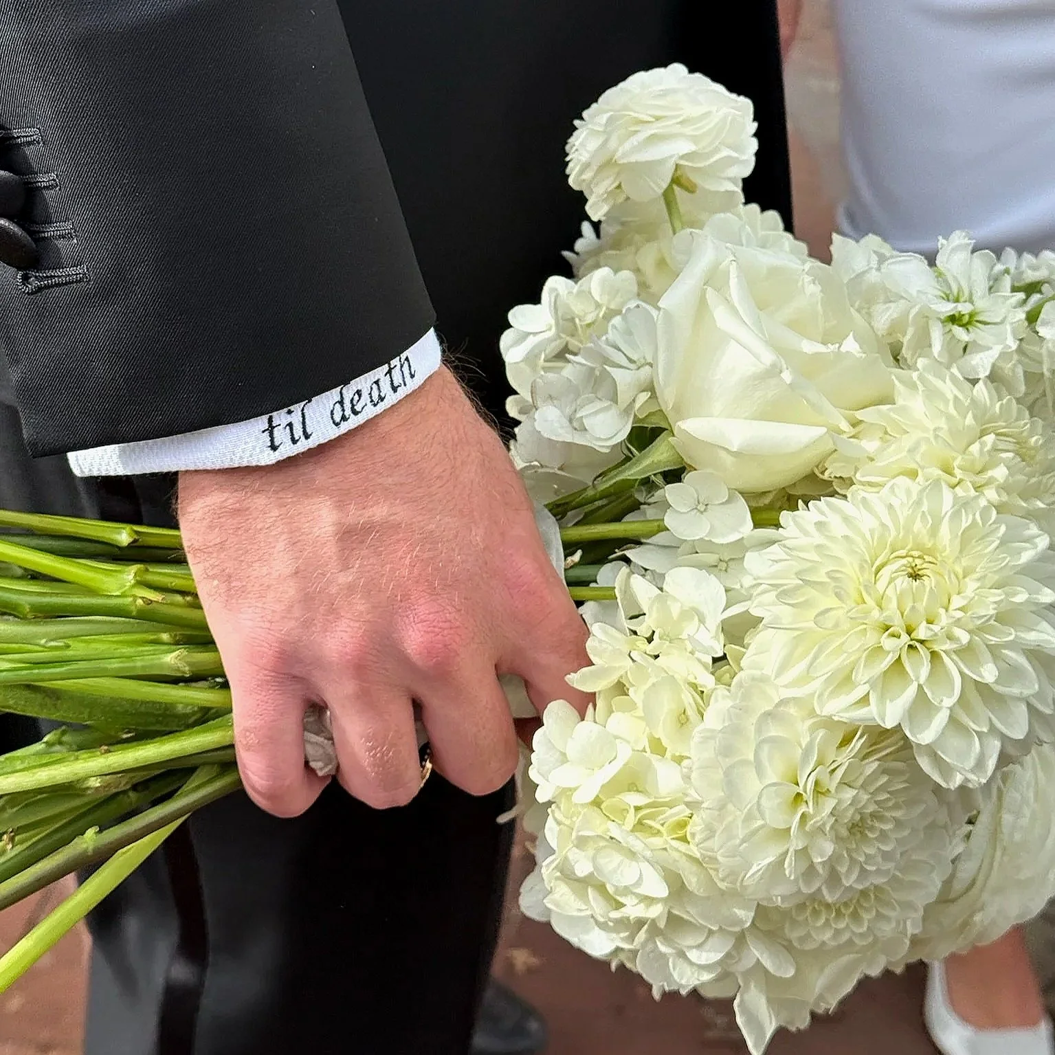 A person in a black suit holding a large bouquet of white flowers, including roses, dahlias, and hydrangeas, with the suit sleeve displaying the phrase 'til death'.