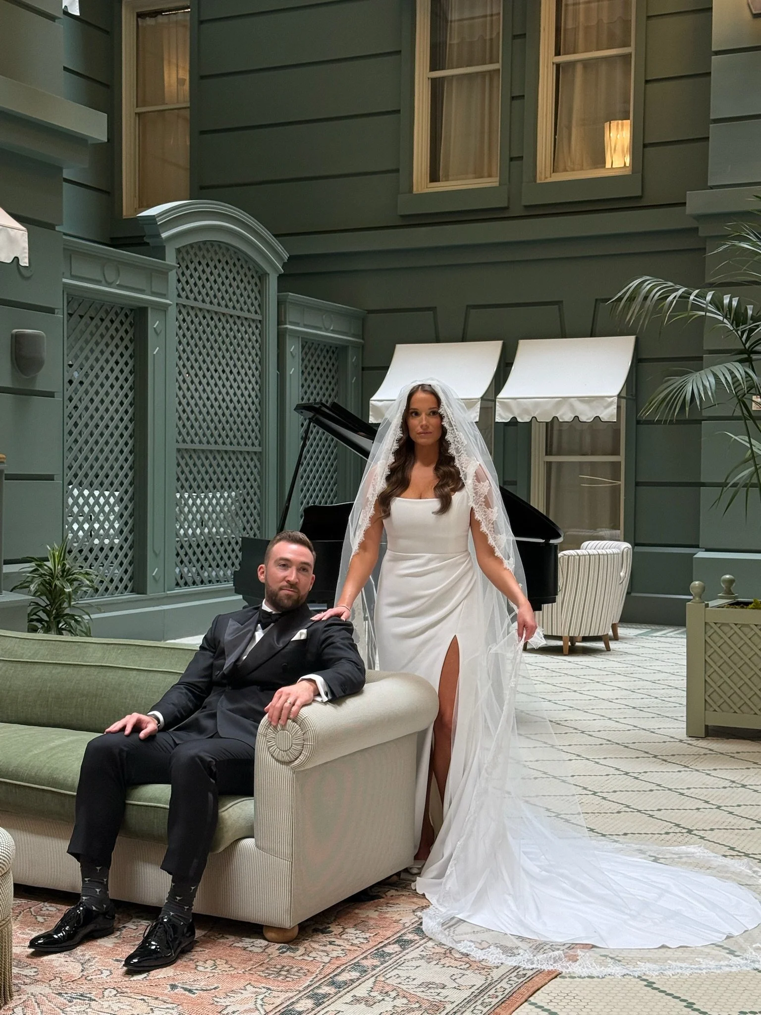 A bride and groom in an elegant, indoor setting. The groom is sitting on a light-colored sofa, dressed in a black tuxedo, while the bride is standing beside him in a white wedding gown with a slit, and a lace veil. A black grand piano is behind them, with large windows and white awnings in the background.