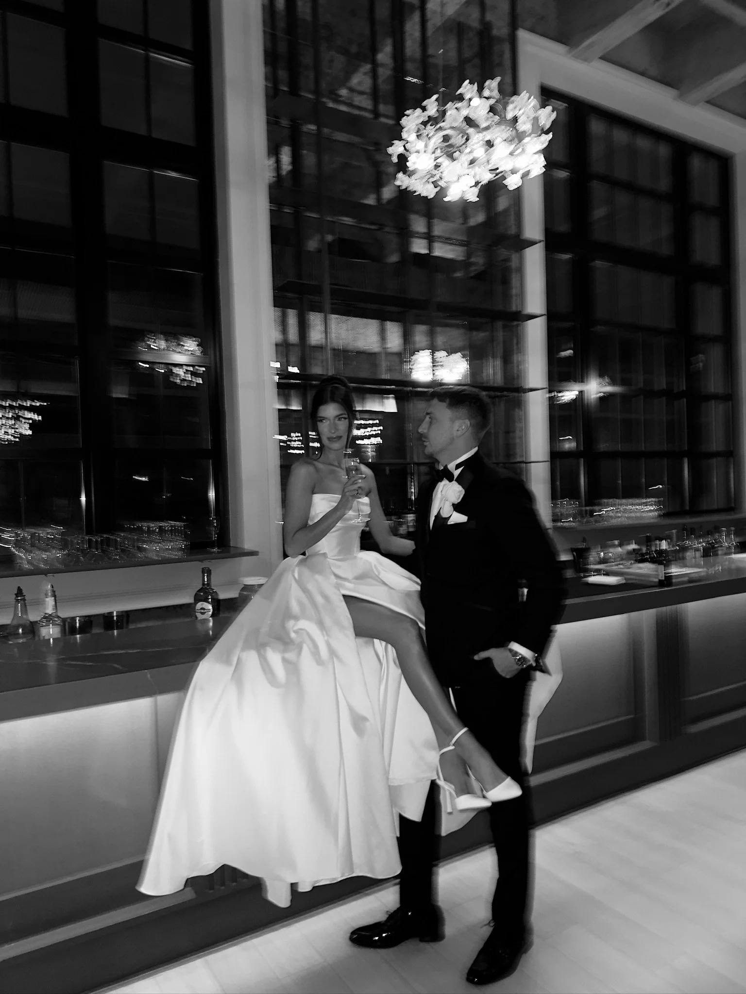 Black and white photo of a woman in a wedding gown sitting on a bar counter, holding a drink, with a man in a tuxedo standing beside her in an elegant indoor setting.