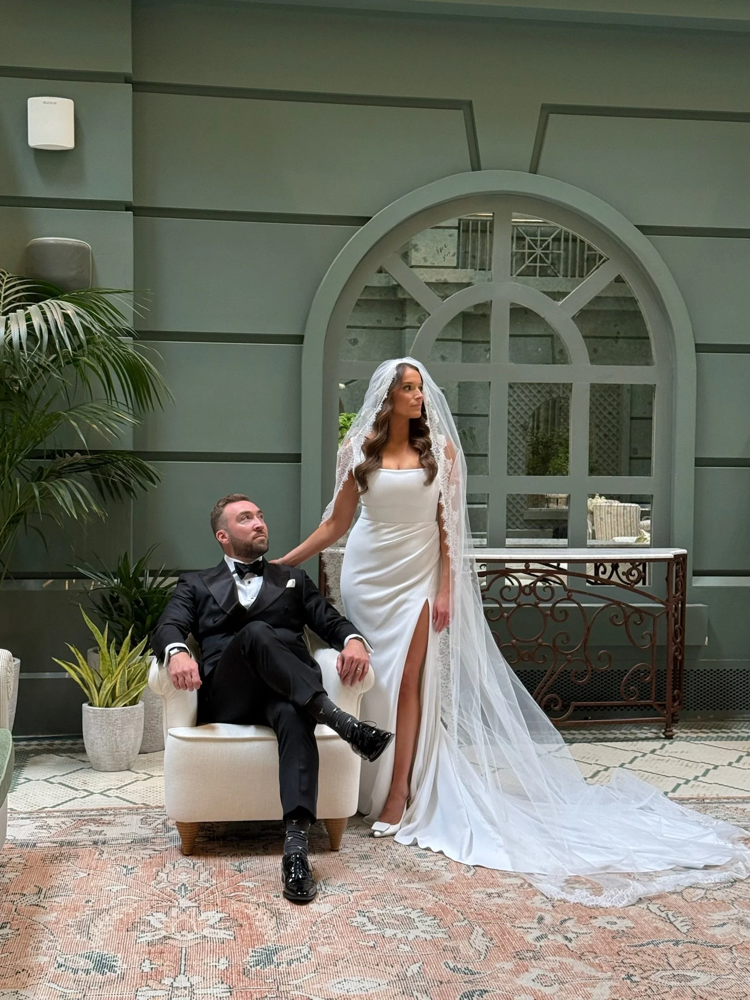 Bride in a white wedding dress and veil standing next to a groom in a tuxedo sitting on a white chair inside a stylish indoor venue with plants and large arched window.