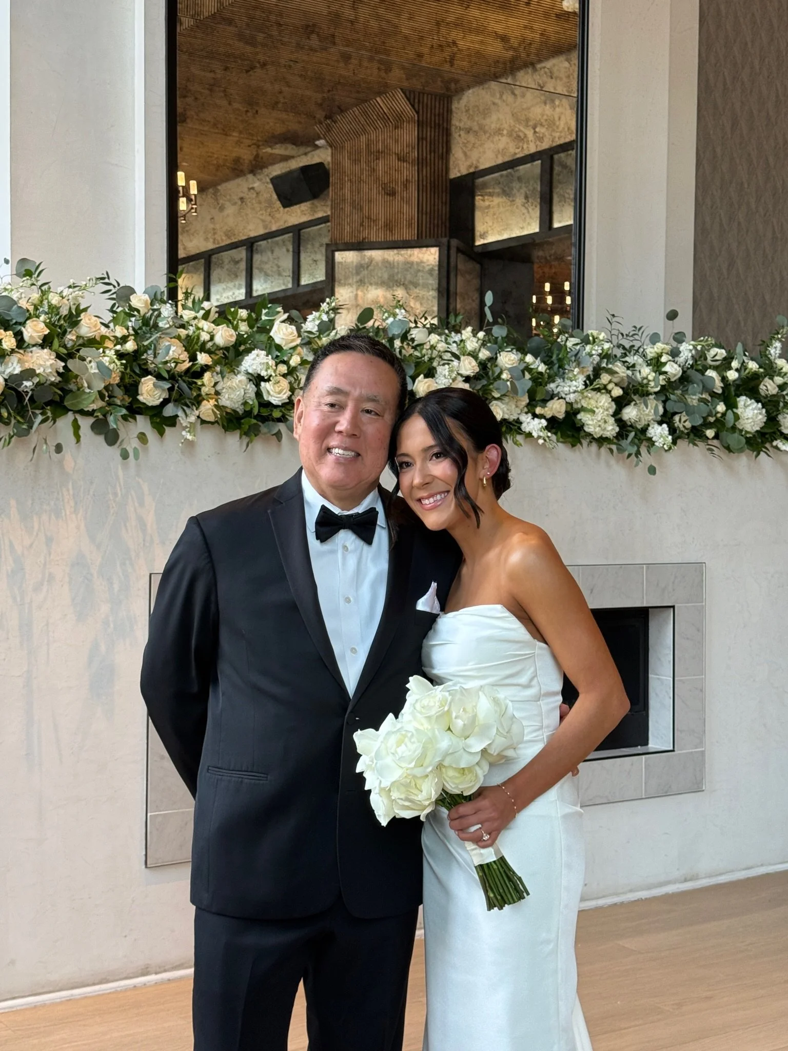 A man in a black tuxedo and a woman in a white wedding dress are standing together, smiling, in front of a wall decorated with a large floral arrangement. The woman is holding a bouquet of white roses.