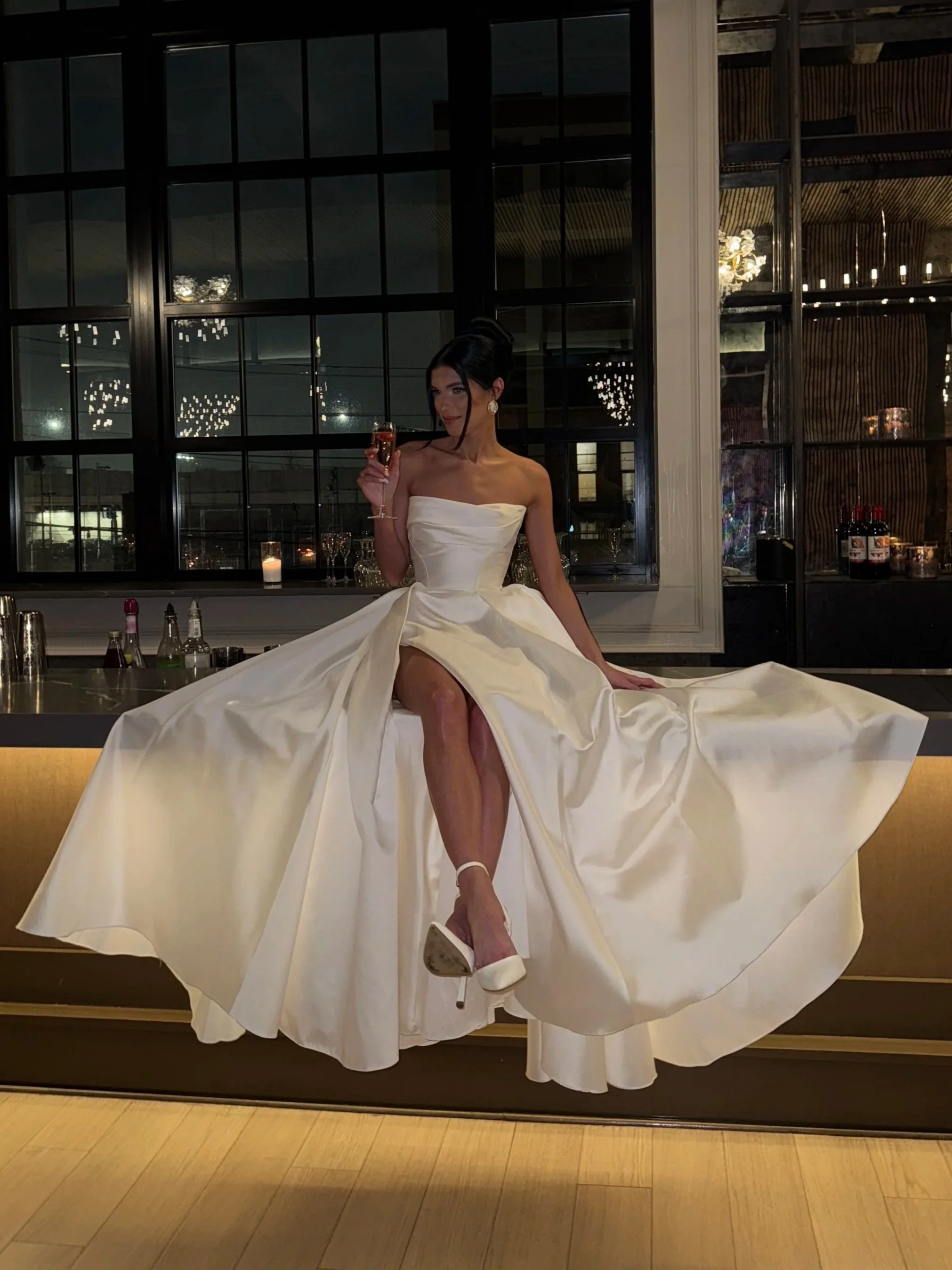 A woman in a strapless white wedding gown sitting on a bar counter at night, holding a glass of champagne, with a large window and elegant chandelier visible in the background.