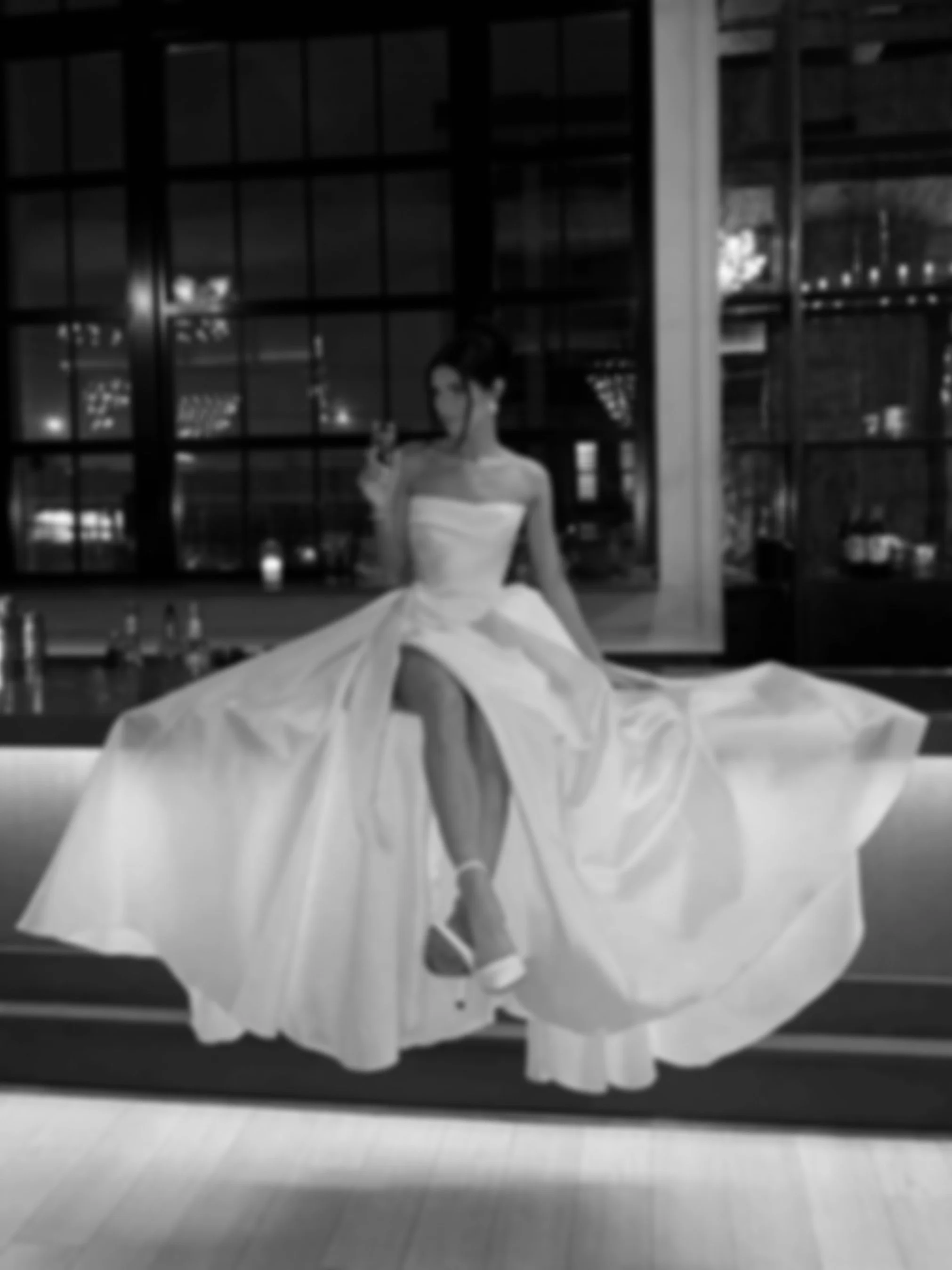 A woman in a wedding dress sitting on a ledge, taking a selfie with her phone in a dimly lit, modern interior with large windows in the background.