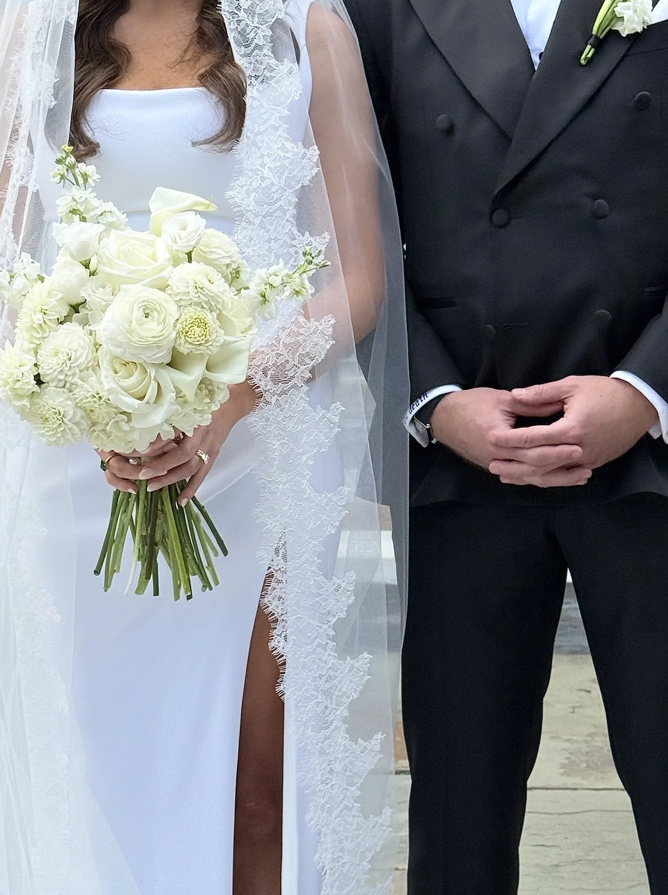 A bride dressed in a white wedding gown holding a bouquet of white flowers standing next to a groom in a dark tuxedo with a white boutonniere.