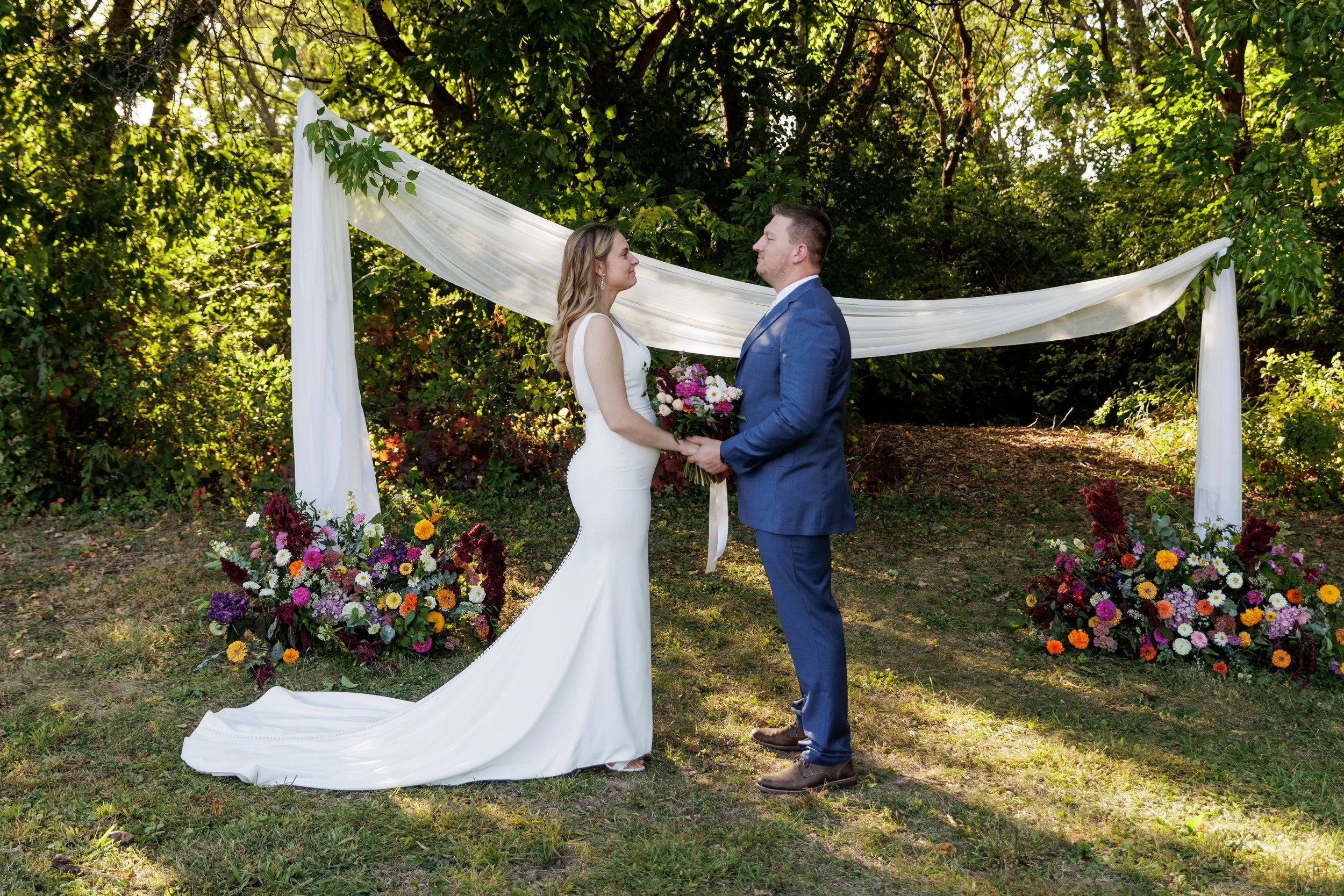 A bride and groom holding hands, exchanging vows outdoors during their wedding ceremony, with greenery and colorful flowers in the background and a white draped fabric arch.