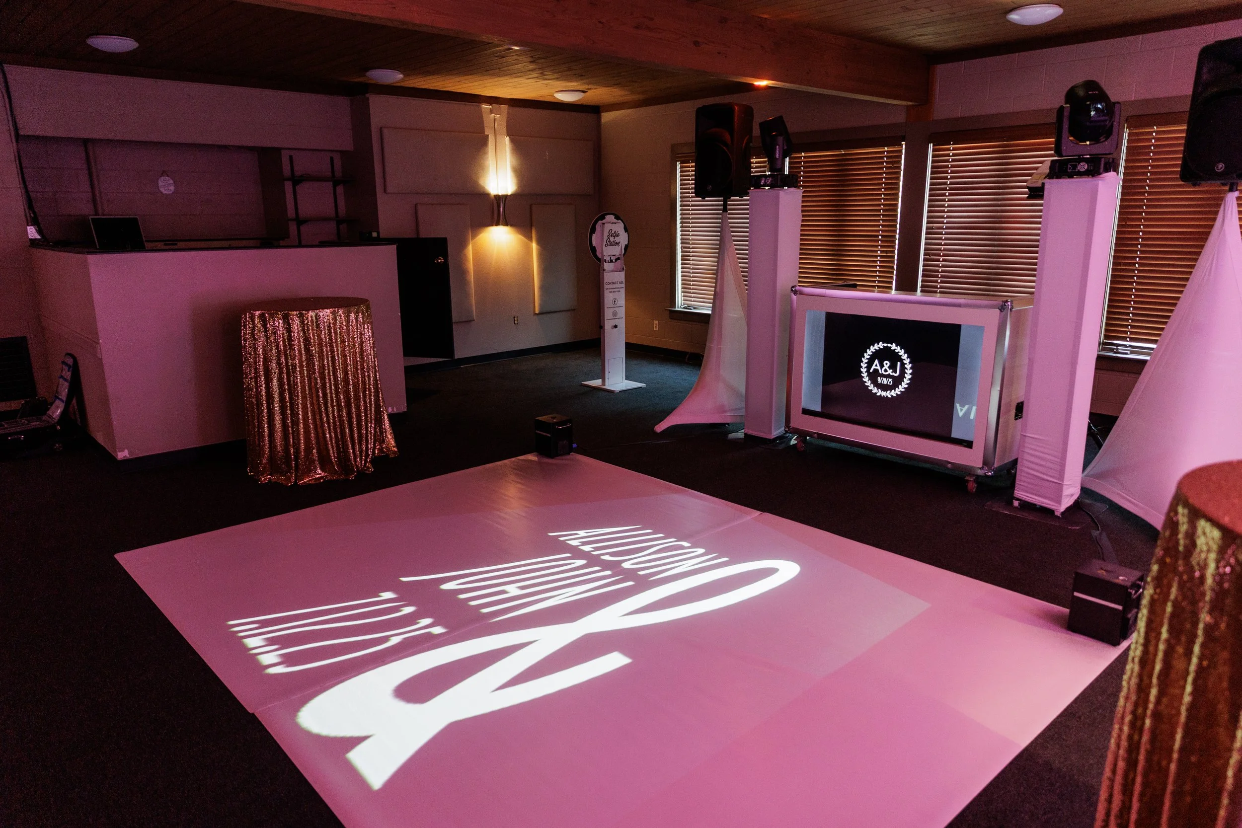 Indoor event space with black flooring and wooden ceiling, set up with DJ equipment, large speakers, and a white-lit floor projection displaying the initials A&J and text. There are windows with closed blinds, a table covered with pink sequined cloth