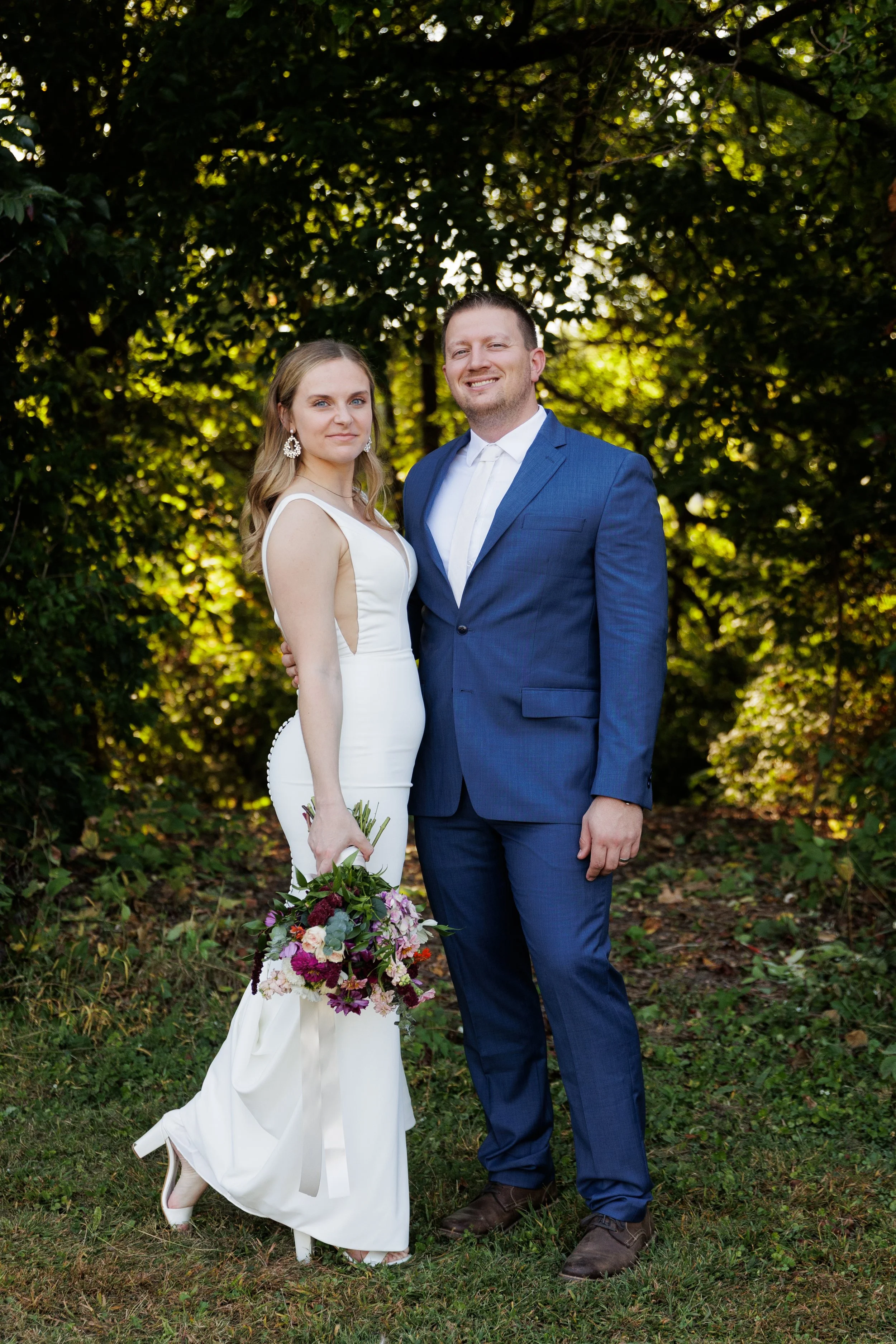 A bride and groom standing outdoors in front of lush green foliage, dressed in wedding attire, with the bride holding a bouquet of flowers.