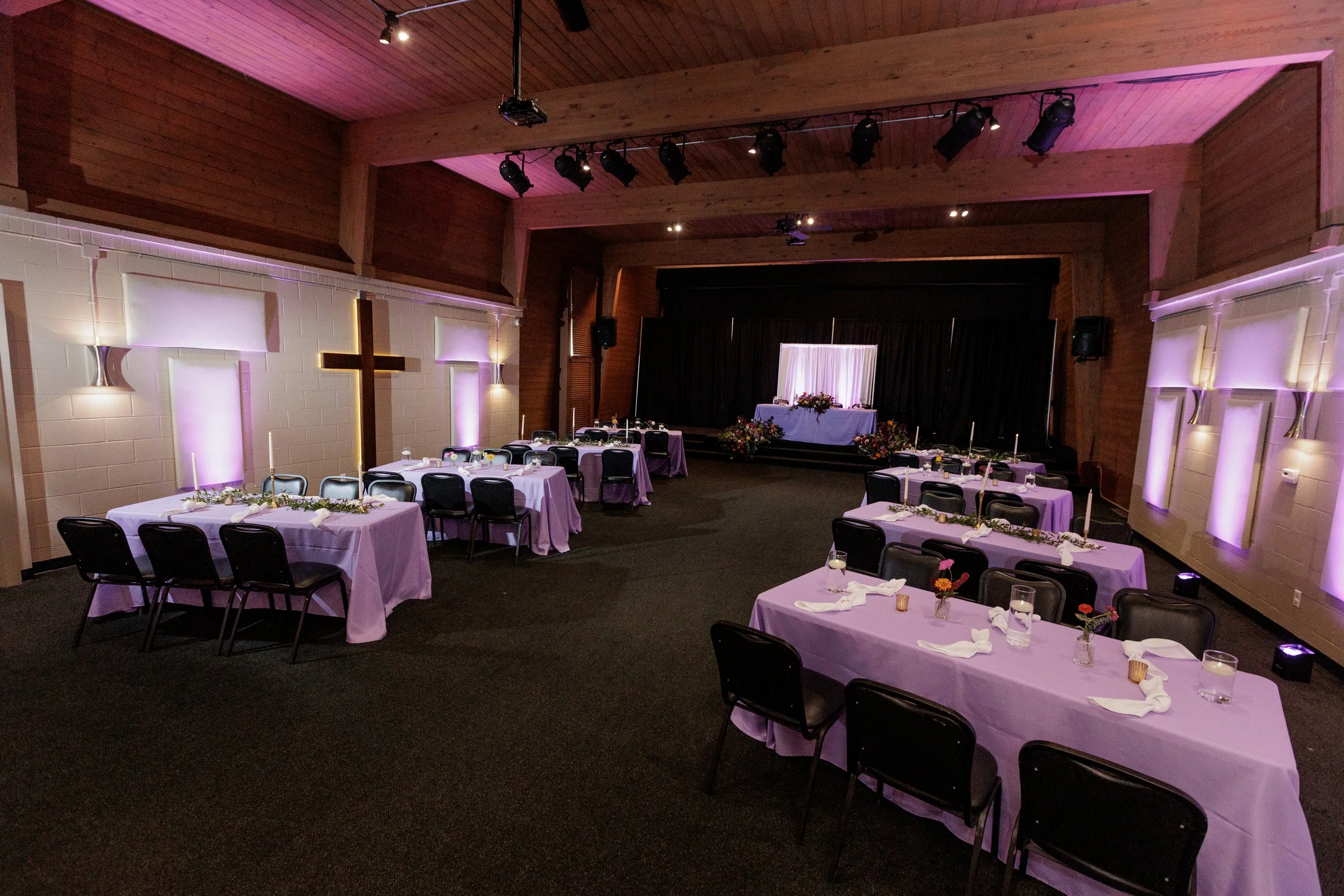 Wedding reception hall with tables covered in purple tablecloths, decorated with small vases of flowers, and tall candles. A raised platform at the front has a table with floral arrangements and a backdrop with curtains, and stage lighting creates a 