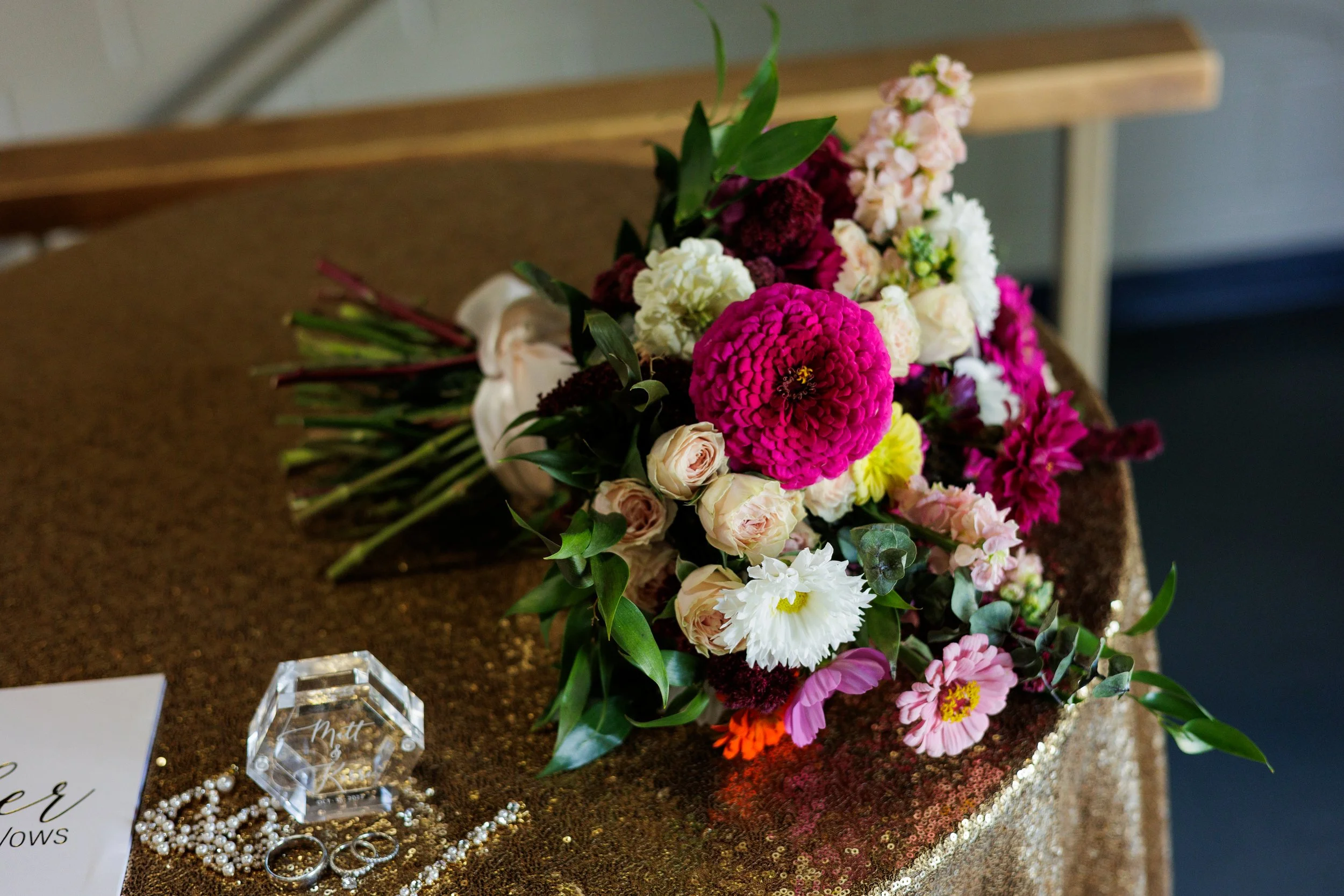 A colorful bouquet of various flowers including pink, white, and purple blooms, resting on a glittery gold table. Nearby are wedding rings, pearl jewelry, and a decorative glass block.
