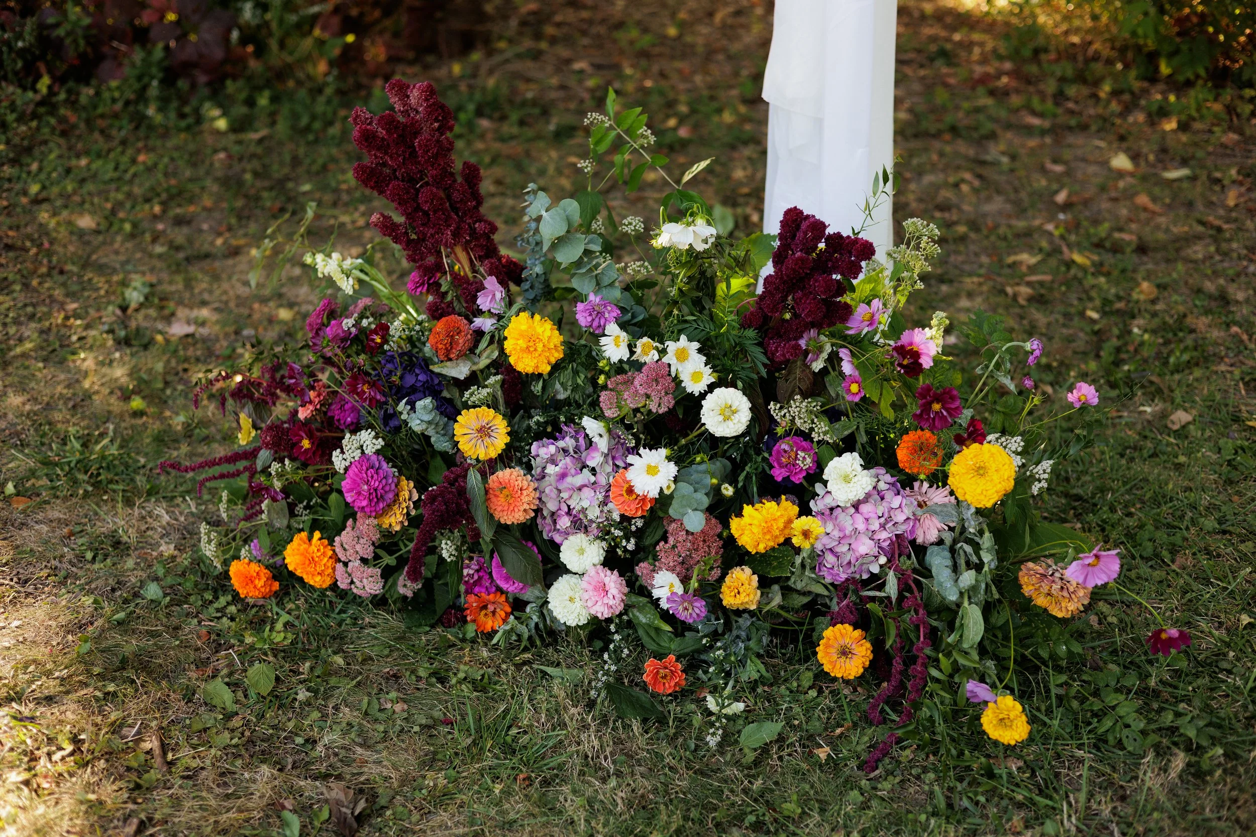 A colorful floral arrangement on the ground, including marigolds, zinnias, hydrangeas, and other assorted flowers, with greenery and some maroon-colored blooms, next to a white post on a grassy, slightly patchy lawn.