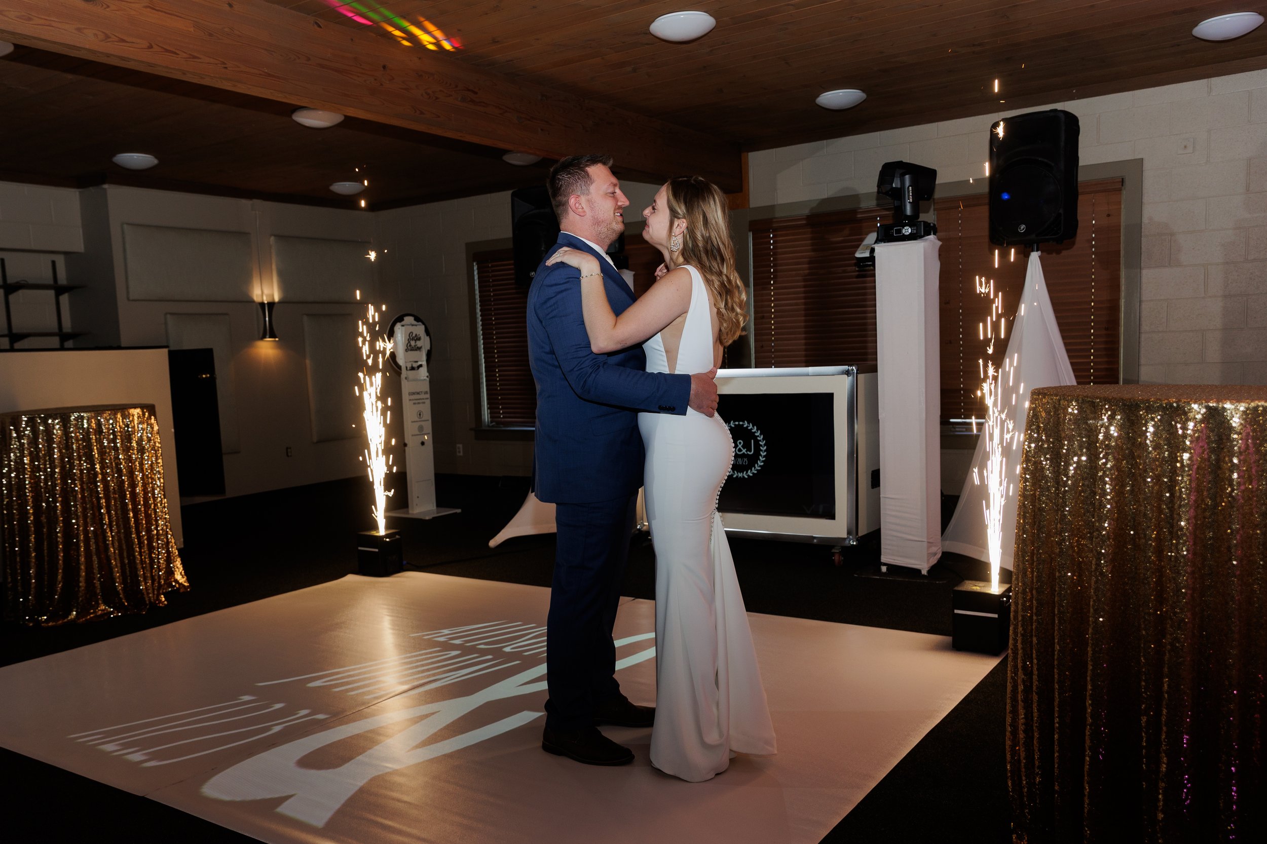 A newlywed couple in wedding attire dancing in a decorated reception hall with sparklers, a gold sequin table, and a DJ setup.